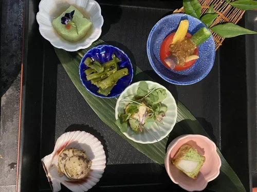 Assorted Japanese desserts and snacks served on small plates and bowls, including matcha-flavored pudding, pickled cucumbers, colorful jelly-like confections, and a green tea cake, all arranged on a large green leaf on a black tray.