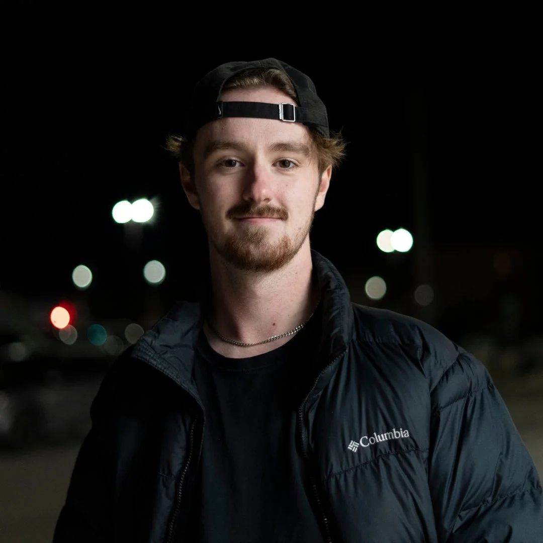 A young man with light brown hair, facial hair, wearing a black baseball cap backwards, a black Columbia jacket, and a silver chain necklace, standing outdoors at night with blurred city lights in the background.