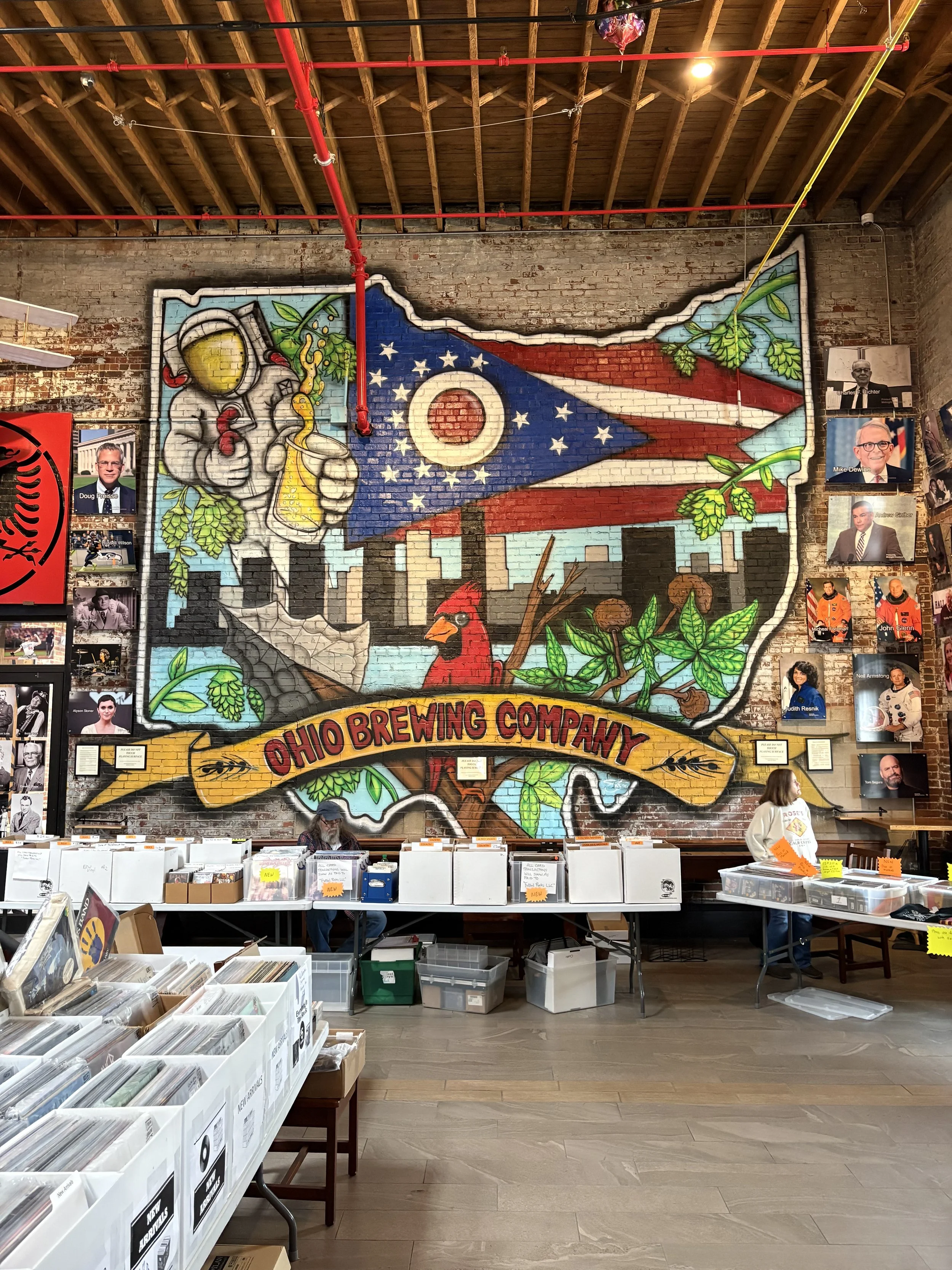 A colorful mural on a brick wall depicts an astronaut holding a beer bottle with hop plant leaves and cones. The mural includes a flag with stars and stripes, and the Ohio Brewing Company sign is visible at the bottom. There are tables with boxes of vinyl records and people browsing. The ceiling is wooden with visible beams and red pipes.