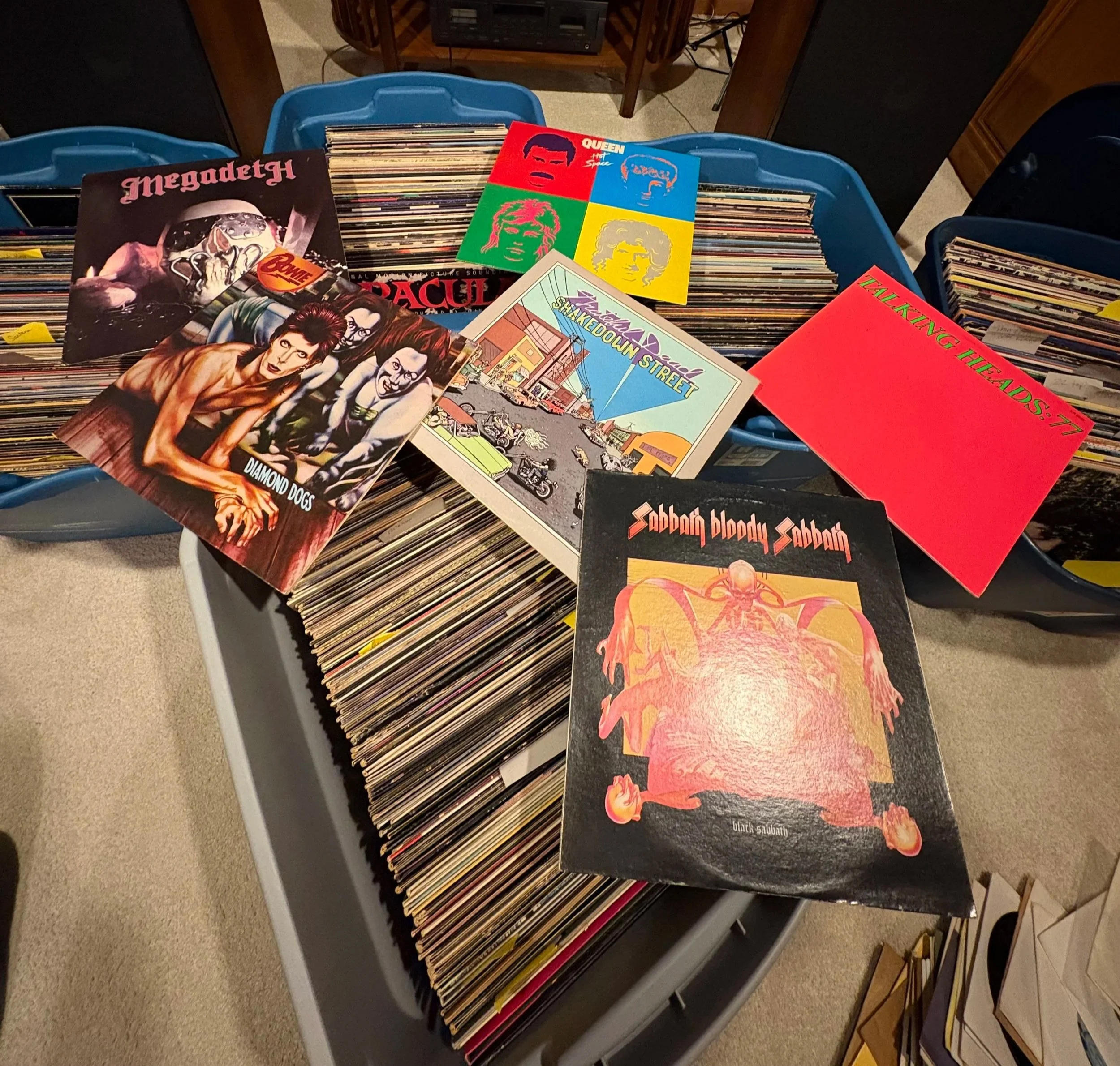 Blue plastic crate filled with vinyl records, with some album covers on top, including Queen's 'The Game' and Megadeth, among other titles.