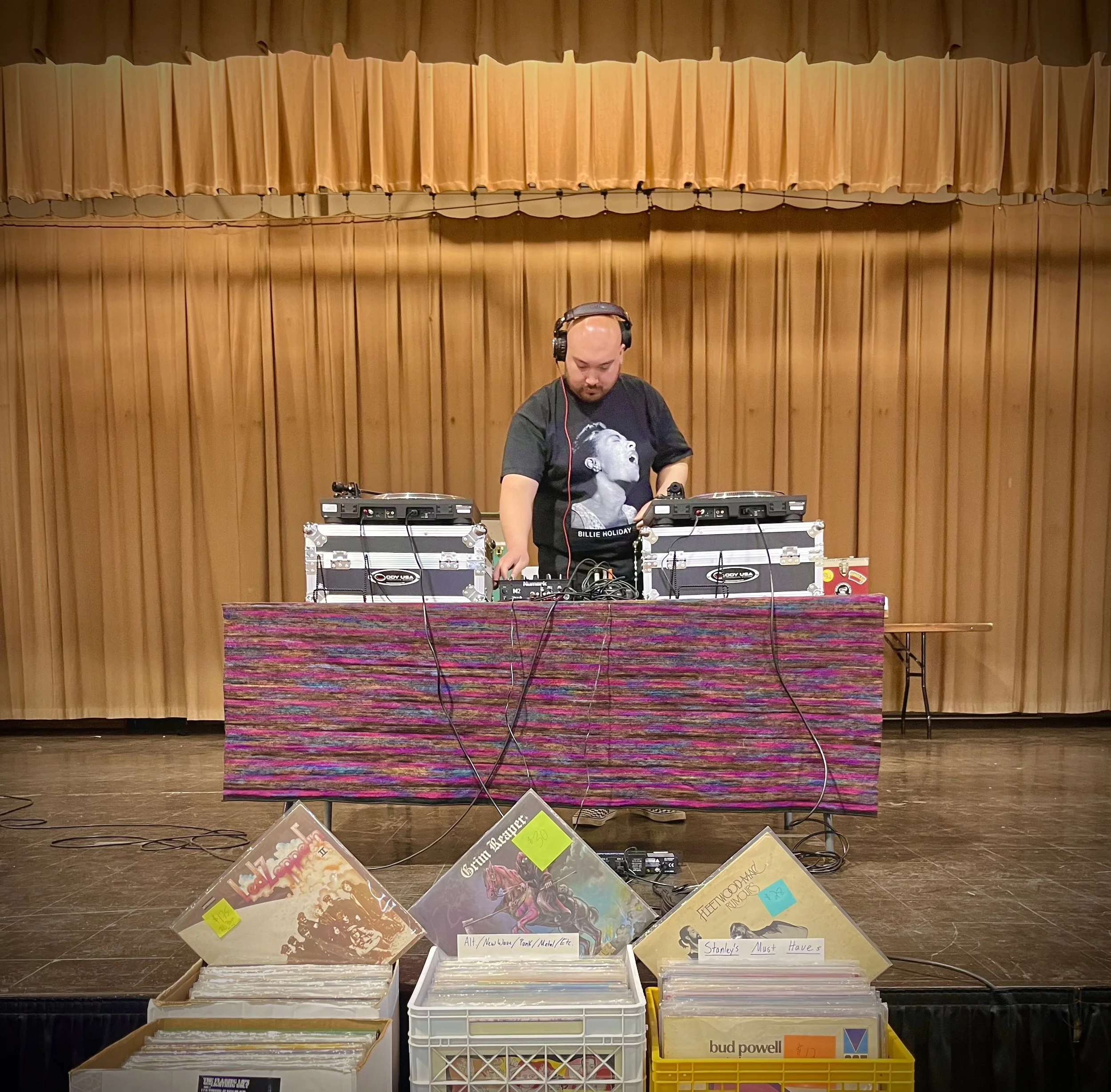 A man wearing headphones and a black T-shirt with a graphic of a woman's face DJing behind a set of turntables in a room with wooden walls and a colorful tablecloth, with displayed vinyl records in front.