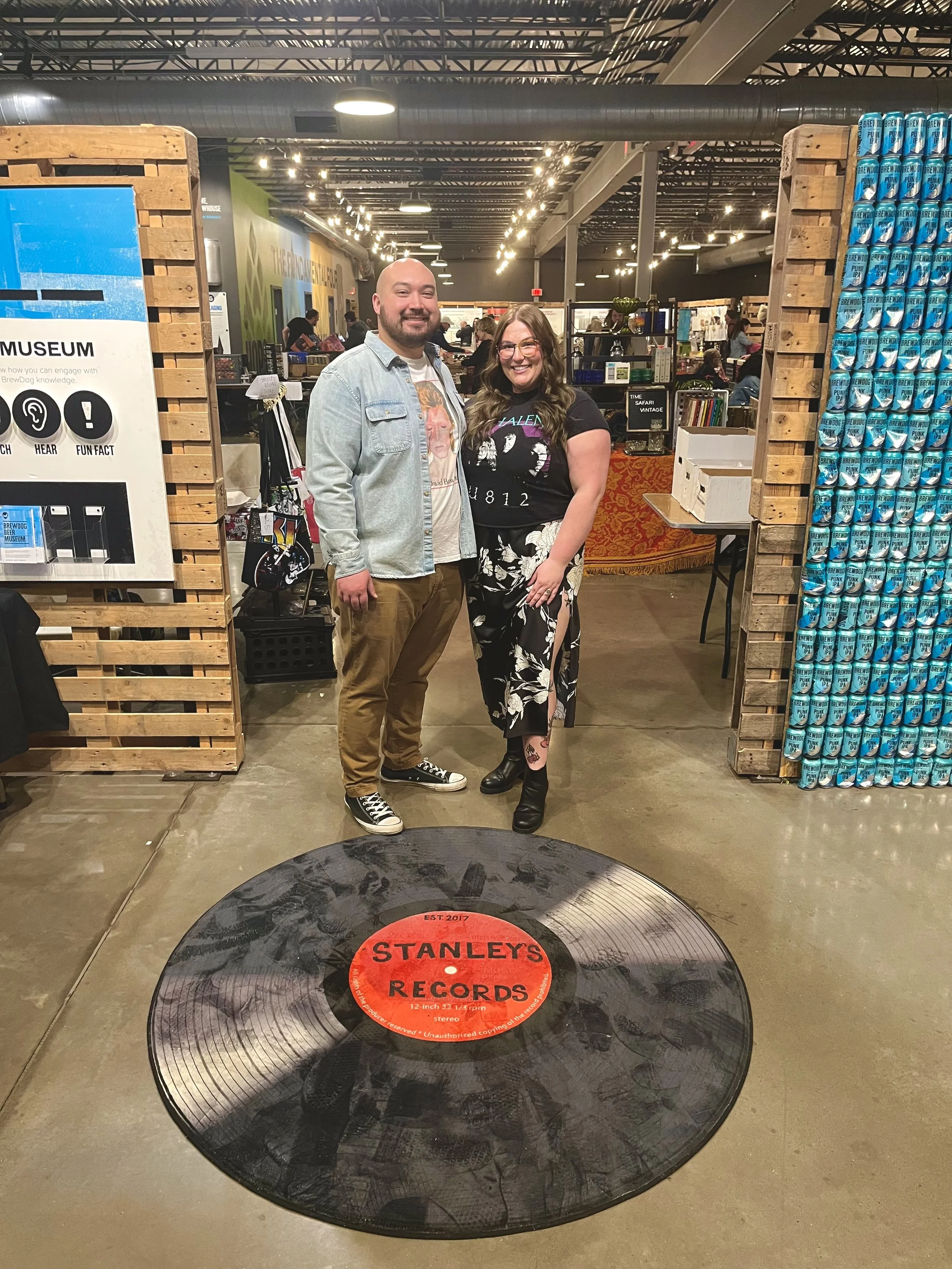 Two people smiling and standing indoors at a store or market, with a large vinyl record on the floor in front of them that has a red sticker reading 'STANLEY'S RECORDS.' The background shows shelves and other customers, with the store's industrial-style ceiling and lighting.