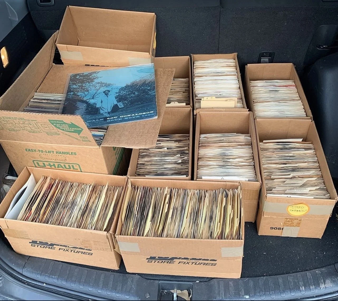 Multiple cardboard boxes filled with manila file folders and documents, some folders are labeled or tabbed. One box has a large blue photo or poster on top. All boxes are placed in the trunk of a vehicle.