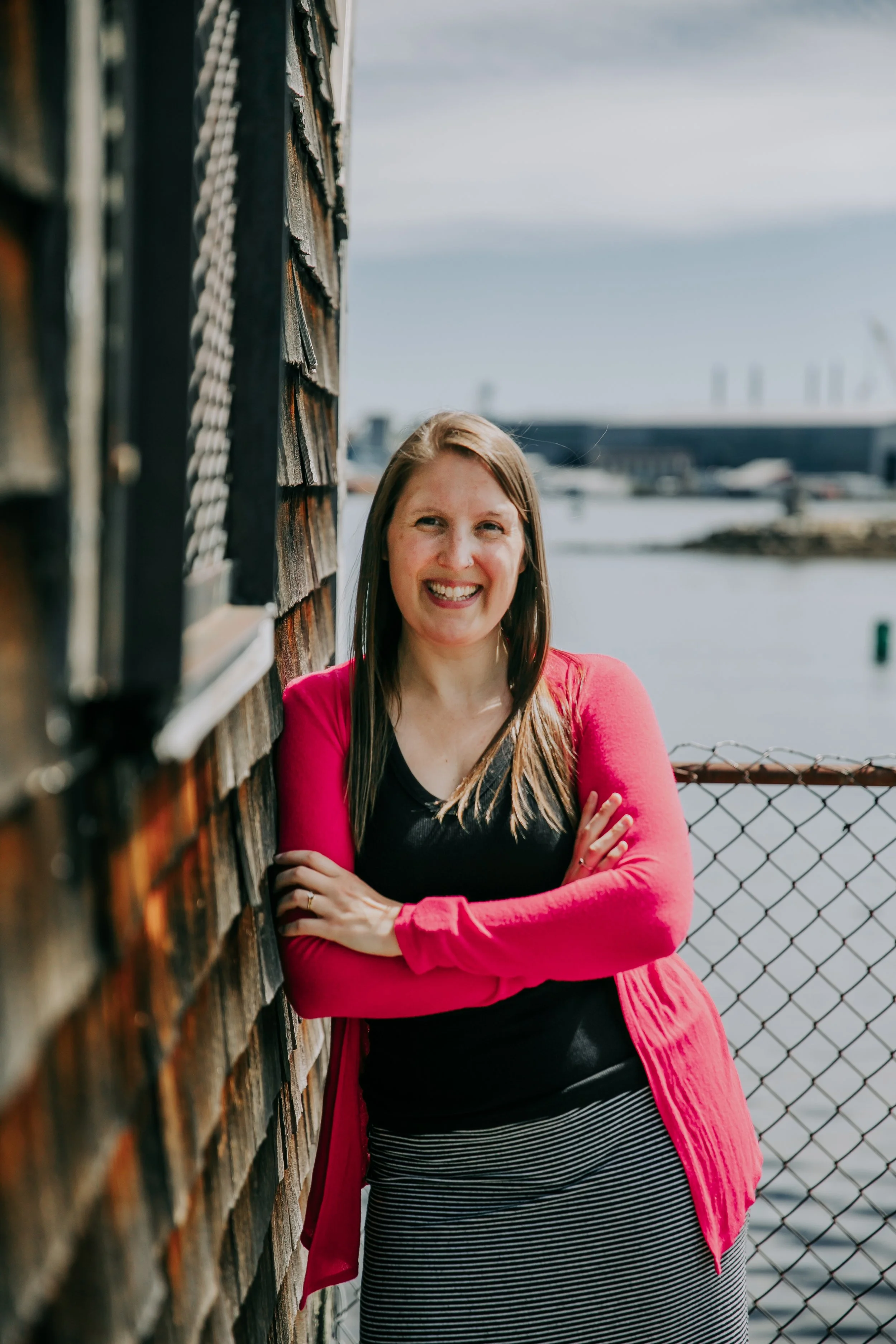 A smiling woman with long hair crossing her arms, wearing a black top, pink cardigan, and black-and-white striped skirt, standing outdoors near a weathered wooden fence and a chain-link fence, with water and industrial buildings in the background.