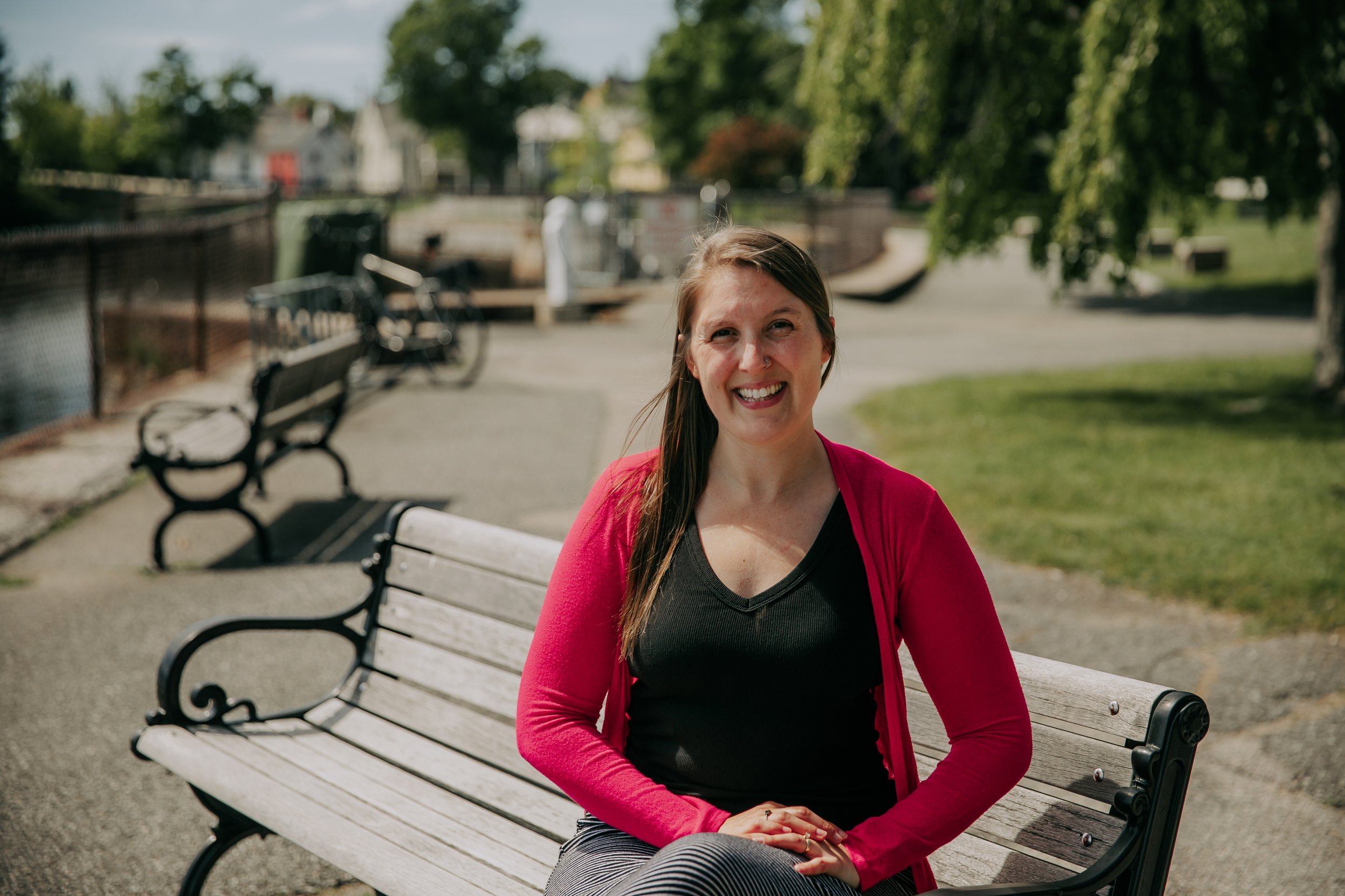 A smiling woman with long brown hair sitting on a park bench, wearing a black top and a red cardigan, with green trees and a pathway in the background.