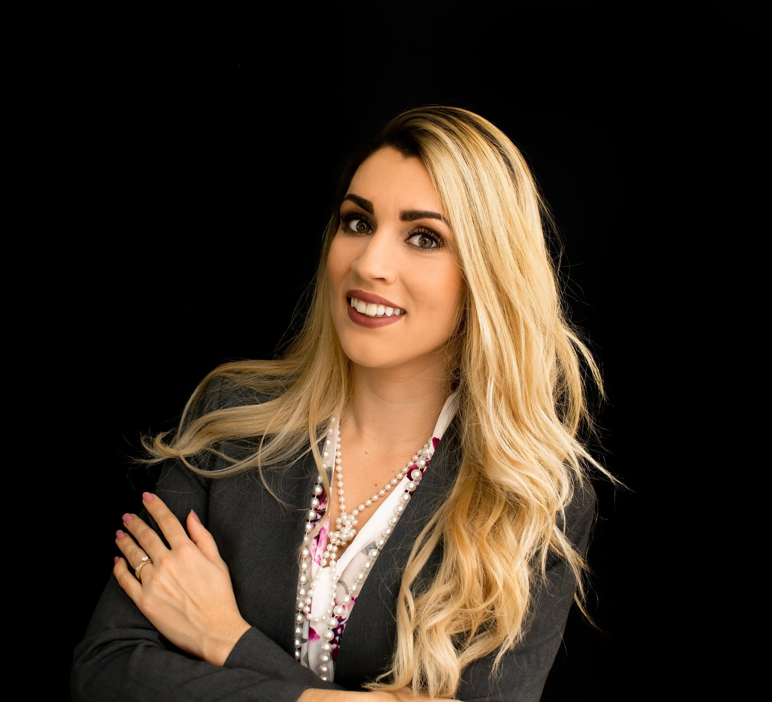 Portrait of a smiling woman with long blond hair, light makeup, wearing a black blazer, pearl necklace, and a white shirt with pink accents, standing against a black background.