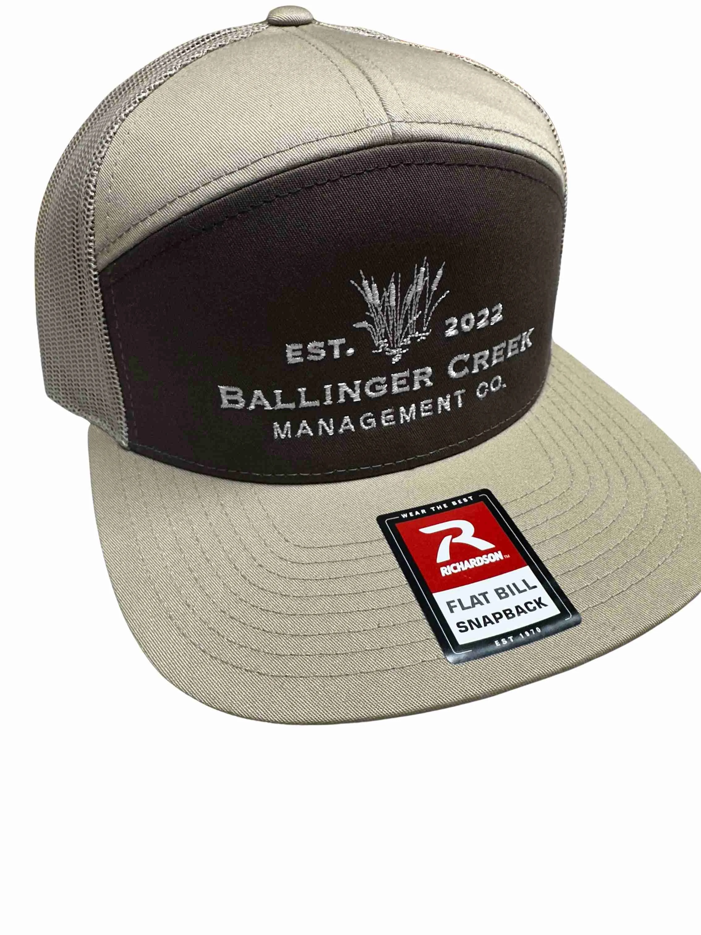 A beige and black snapback cap with embroidered logo and text on the front panel, and a sticker on the brim.