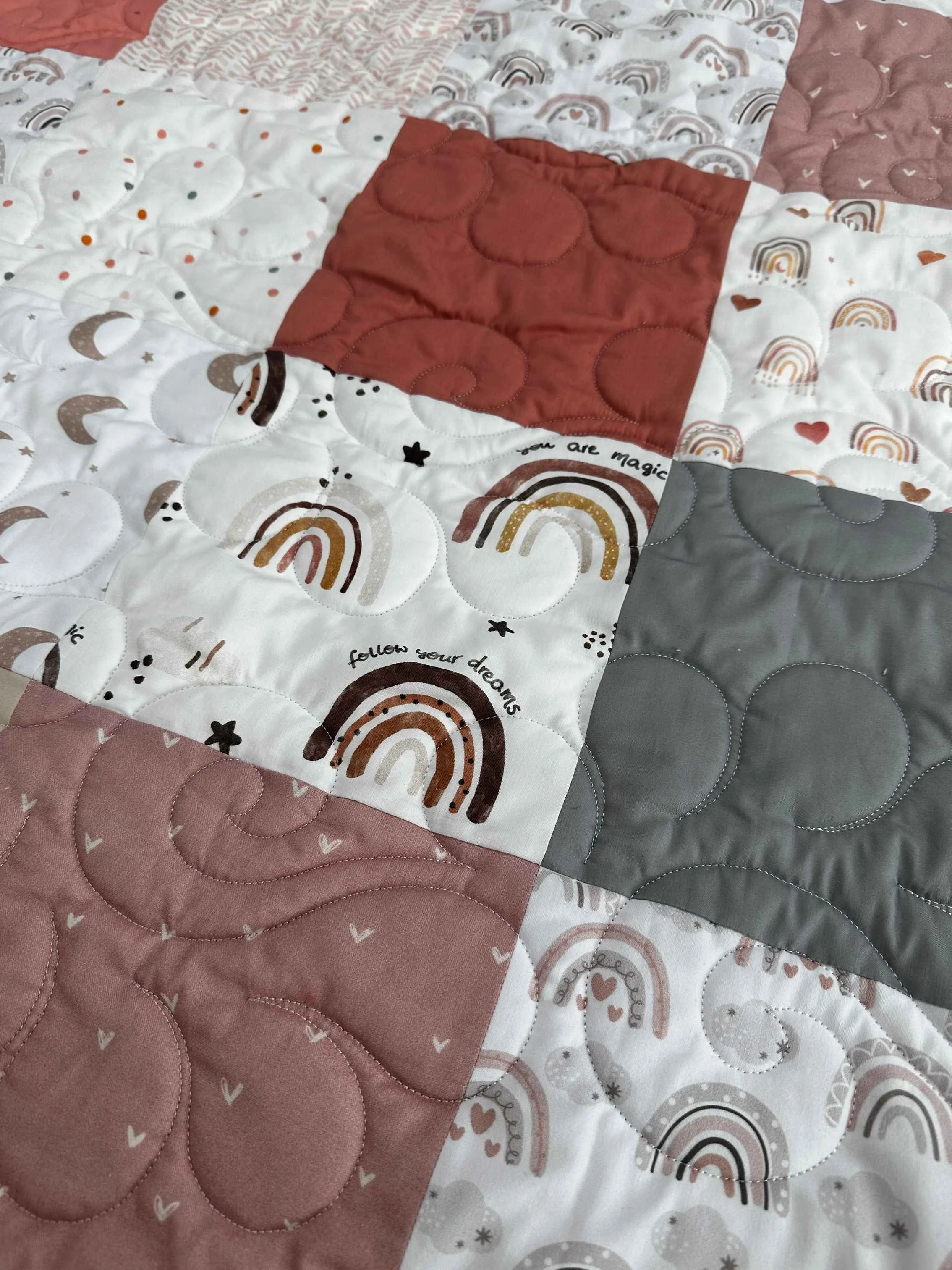 Close-up of a quilt with pink, gray, and white fabric squares, some featuring rainbows, moons, and hearts, with various stitched patterns.