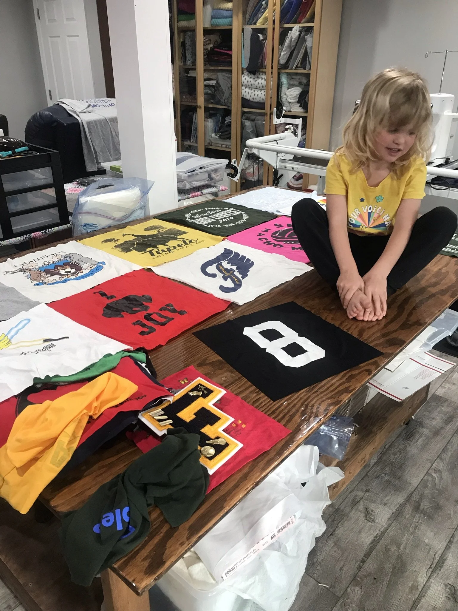 A wooden table displaying various colorful iron-on patches, t-shirts, and clothing items. A young girl with blonde hair, wearing a yellow T-shirt, is sitting on the table with her legs crossed, in a room with a bookshelf and storage shelves in the background.