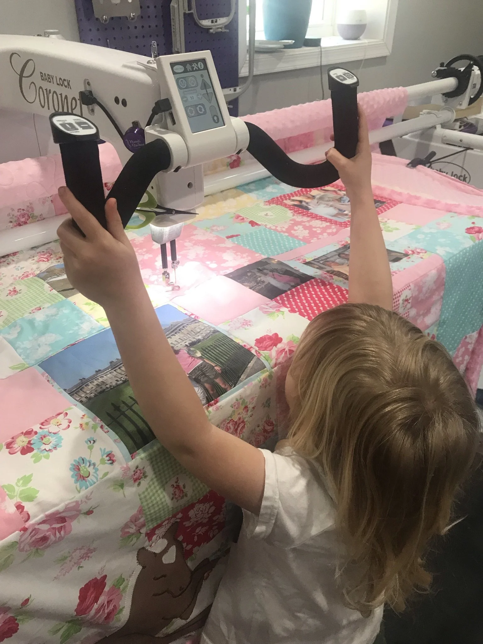 A child with long blonde hair, wearing a white shirt, is reaching out and touching a medical robotic device with a touchscreen display. The device has a white body with black arms, and the background shows a colorful quilted bedspread with various patterns and pictures.