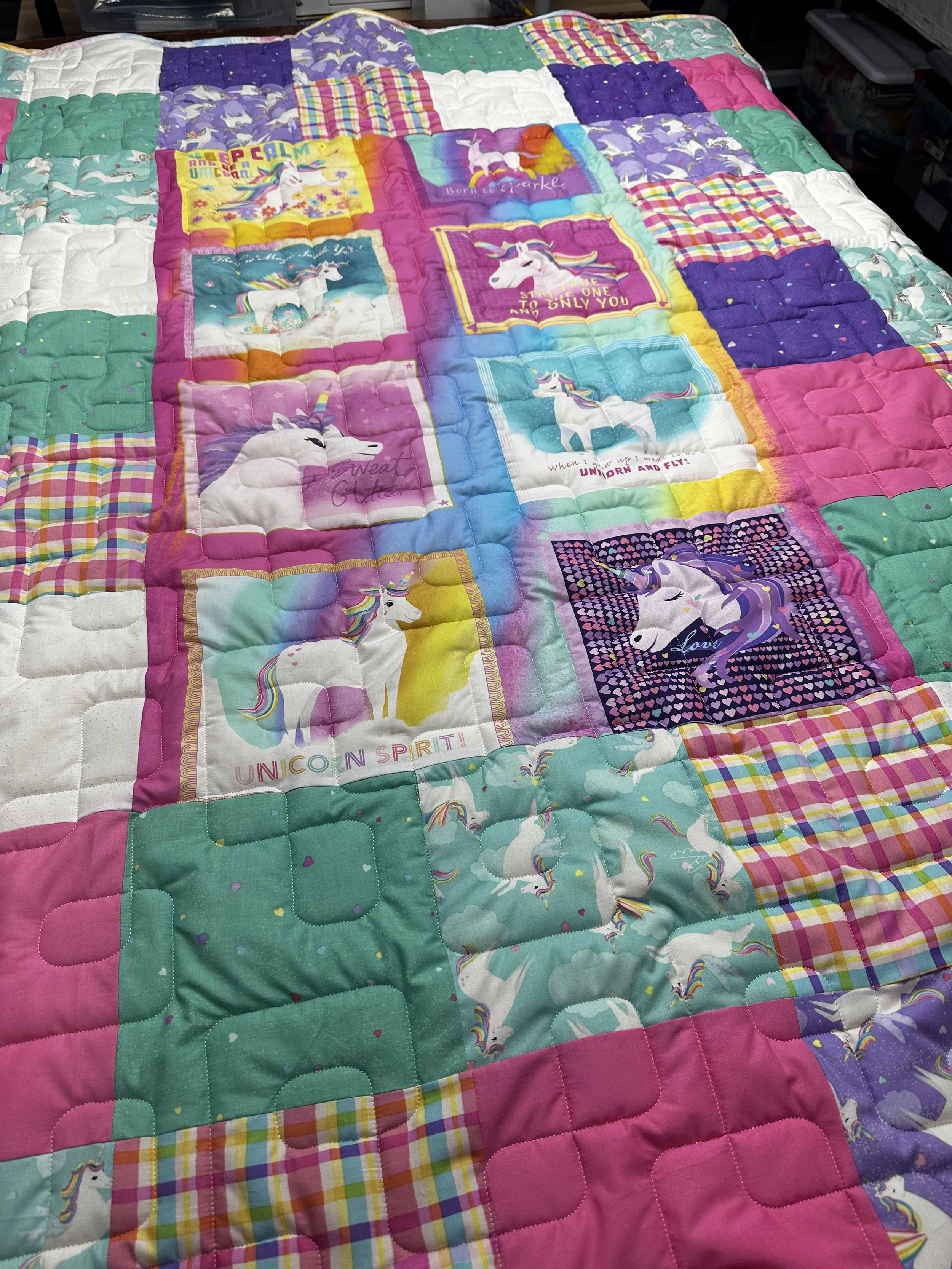 Colorful patchwork quilt featuring various unicorn-themed fabric squares, including images of unicorns, rainbows, and phrases like 'Unicorn Spirit' and 'Love', with a pink, purple, teal, and checkered border.