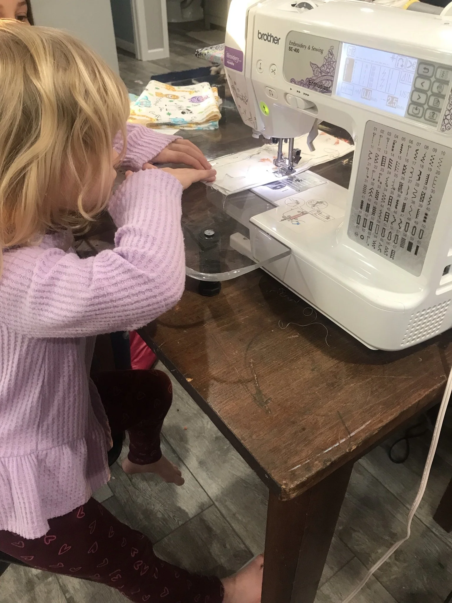 A young girl with blonde hair, wearing a pink sweater and maroon pants with a heart pattern, working on a sewing project on a wooden table. She is using a Brother embroidery and sewing machine. There are fabric pieces and patterns on the table.