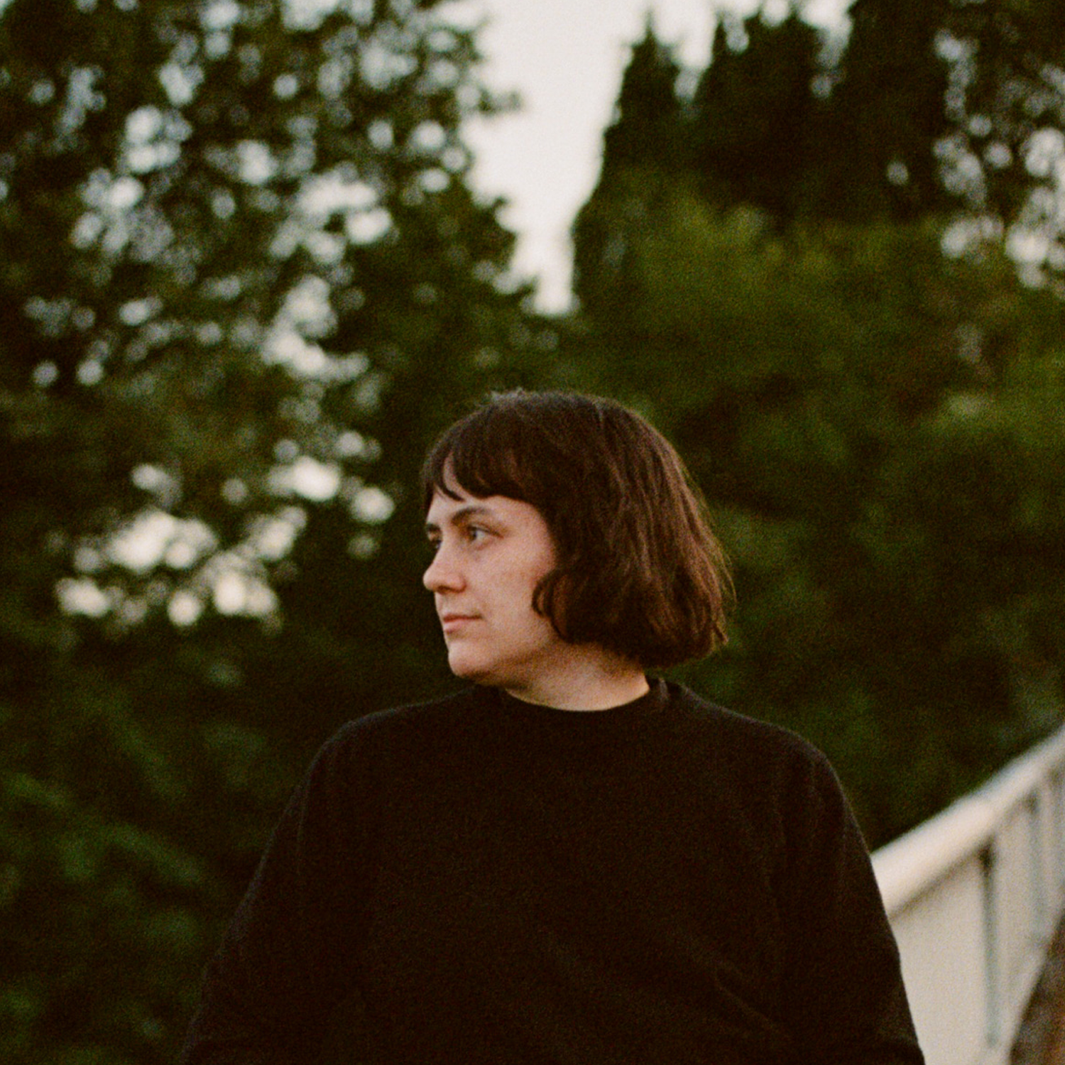 Photo of Madeleine Jubilee Saito, a white woman in her 30s with dark brown hair styled in a bob with bangs. She stands on a bridge in front of a dark forest at dusk.