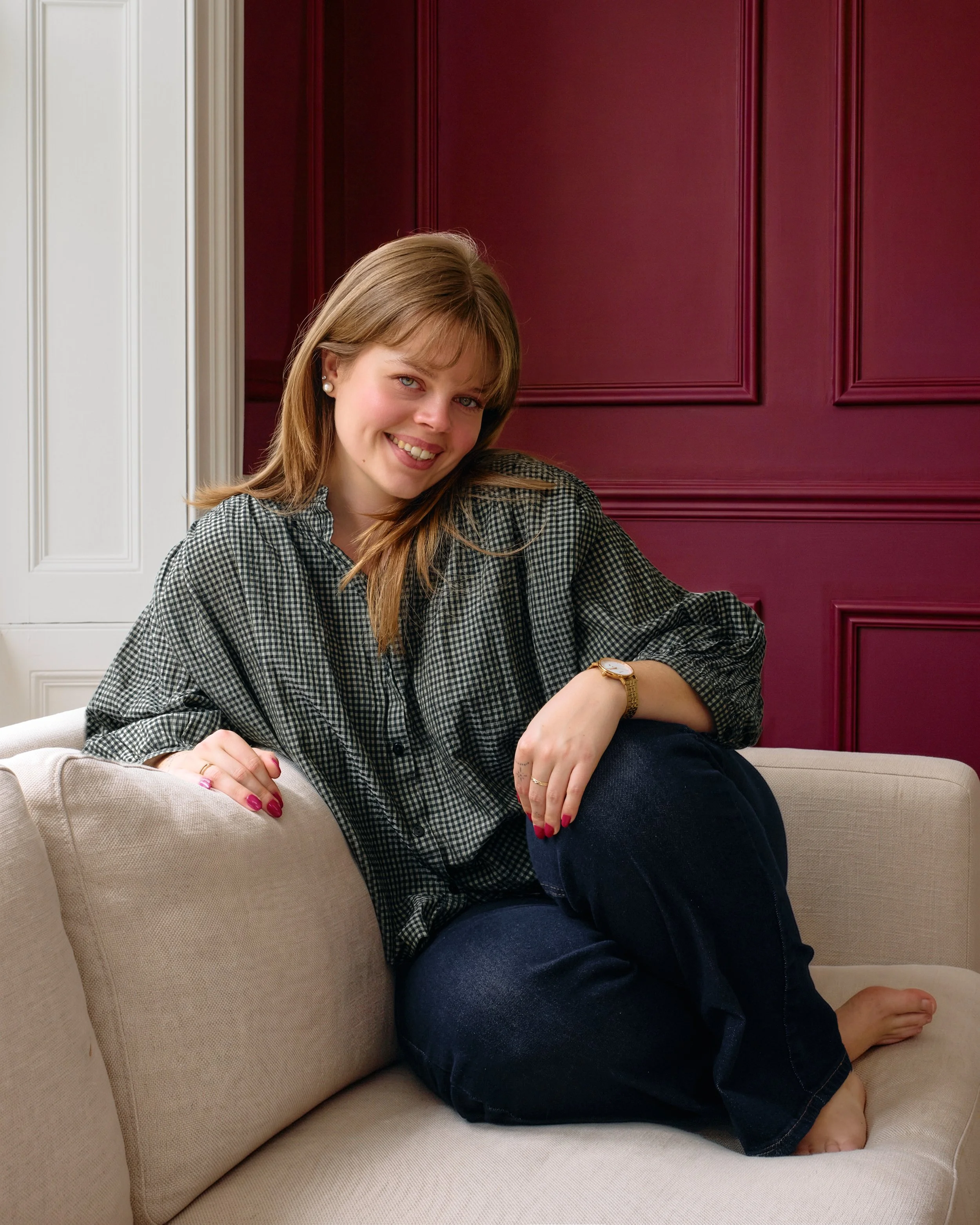 Femme avec cheveux longs, portant un bracelet doré, assise sur un canapé beige, avec un fond de mur rouge.