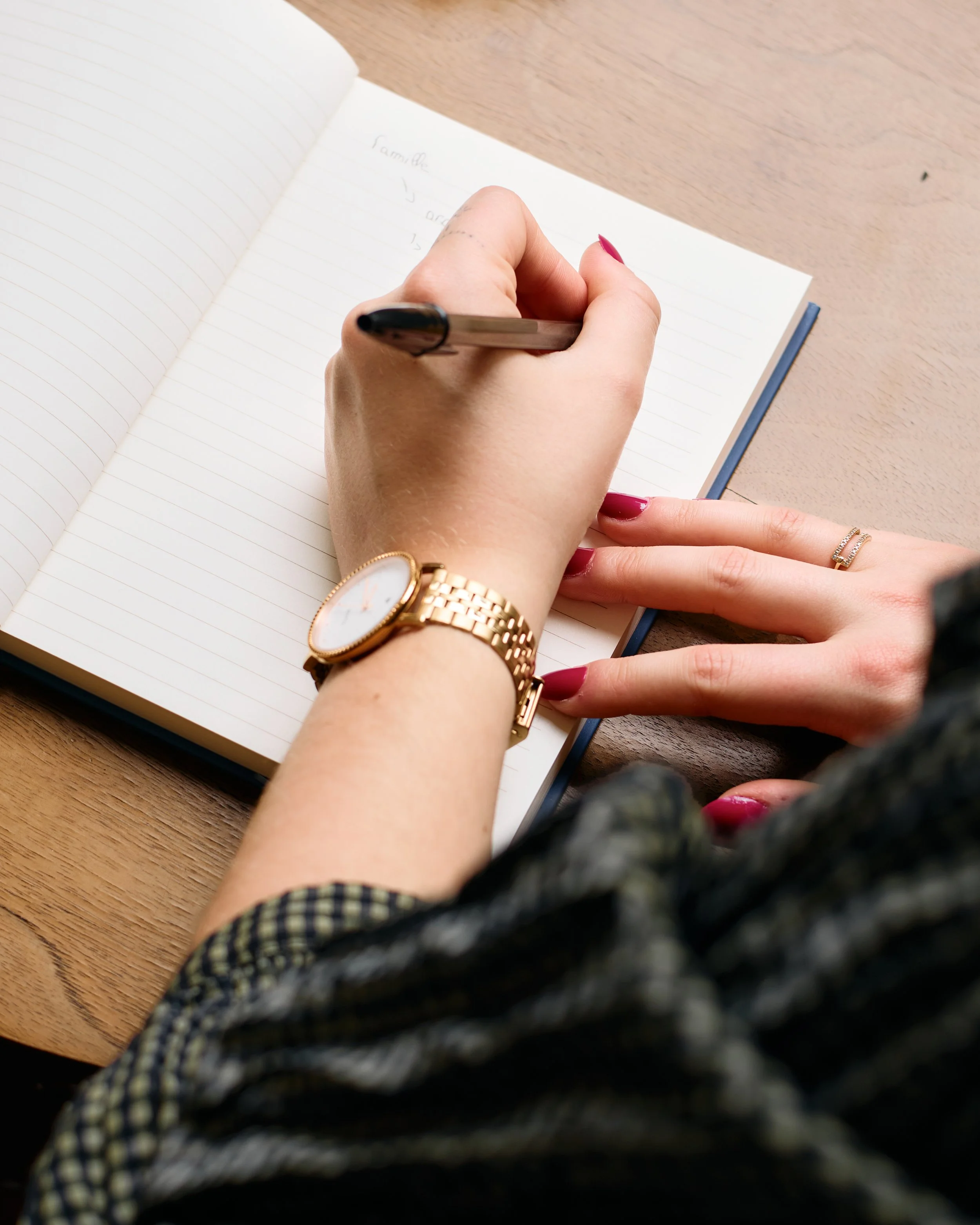 Une personne écrit dans un carnet avec un stylo noir, portant une montre dorée et des bagues, vu de près sur un bureau en bois.