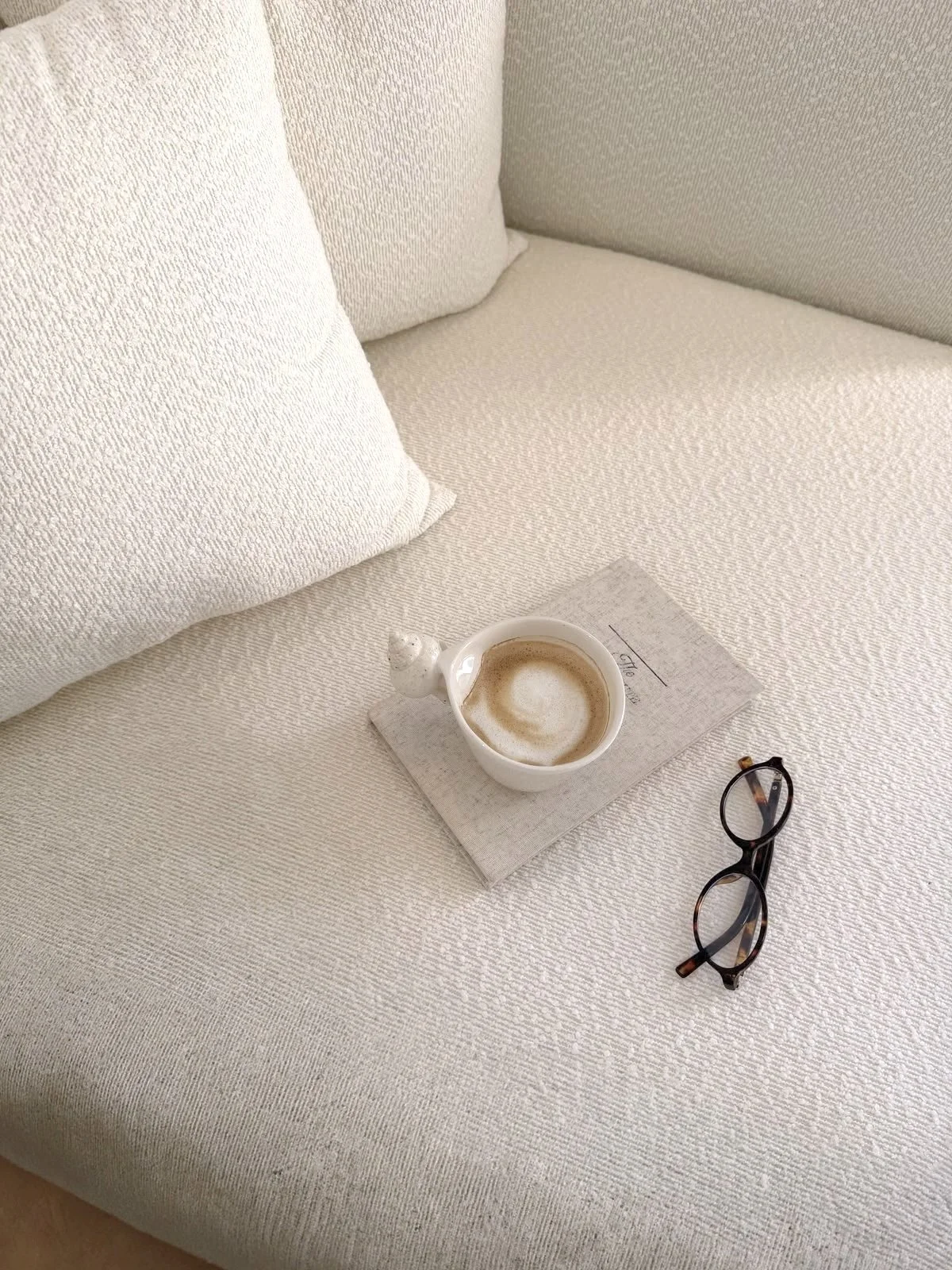 Une tasse de café sur une nappe blanche, accompagnée de lunettes et d'un journal sur un canapé ou lit en tissu blanc