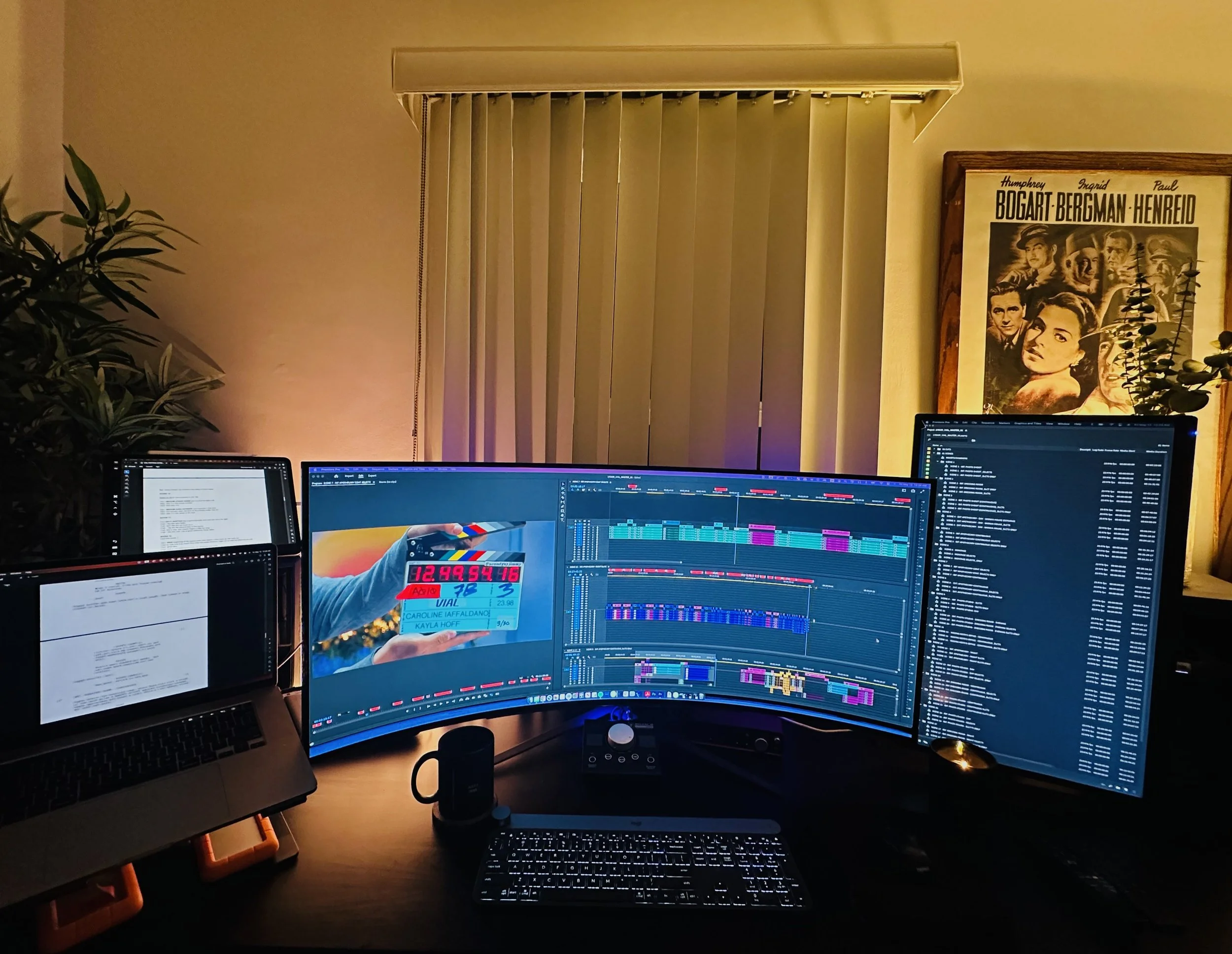 Home office workspace with multiple monitors displaying audio editing software, a laptop with a document open, a keyboard, and a plant in the background. A framed poster of a classic film scene is on the wall, and vertical blinds cover a window.