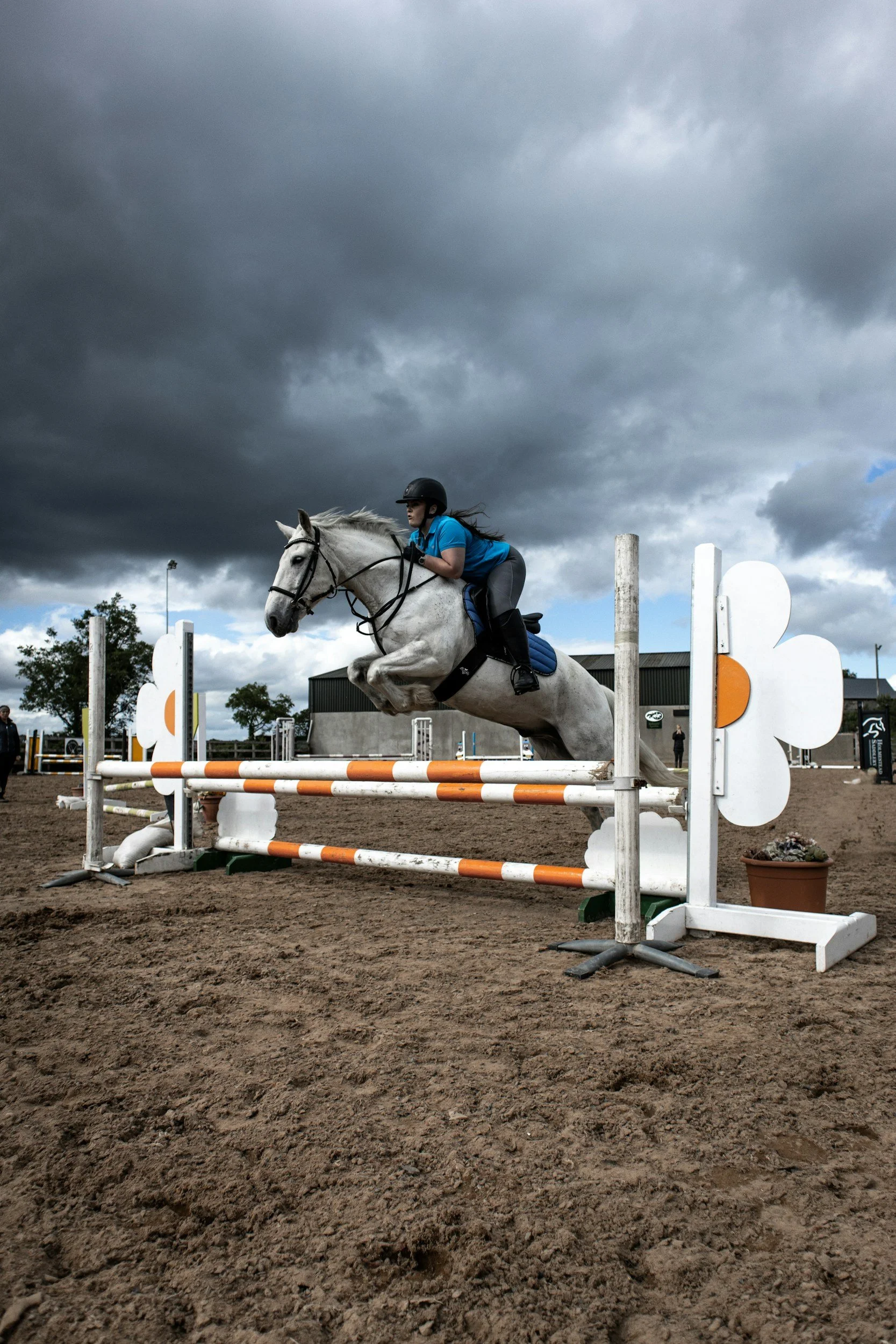 A woman riding a gray horse over a show jumping obstacle outdoors on a cloudy day.