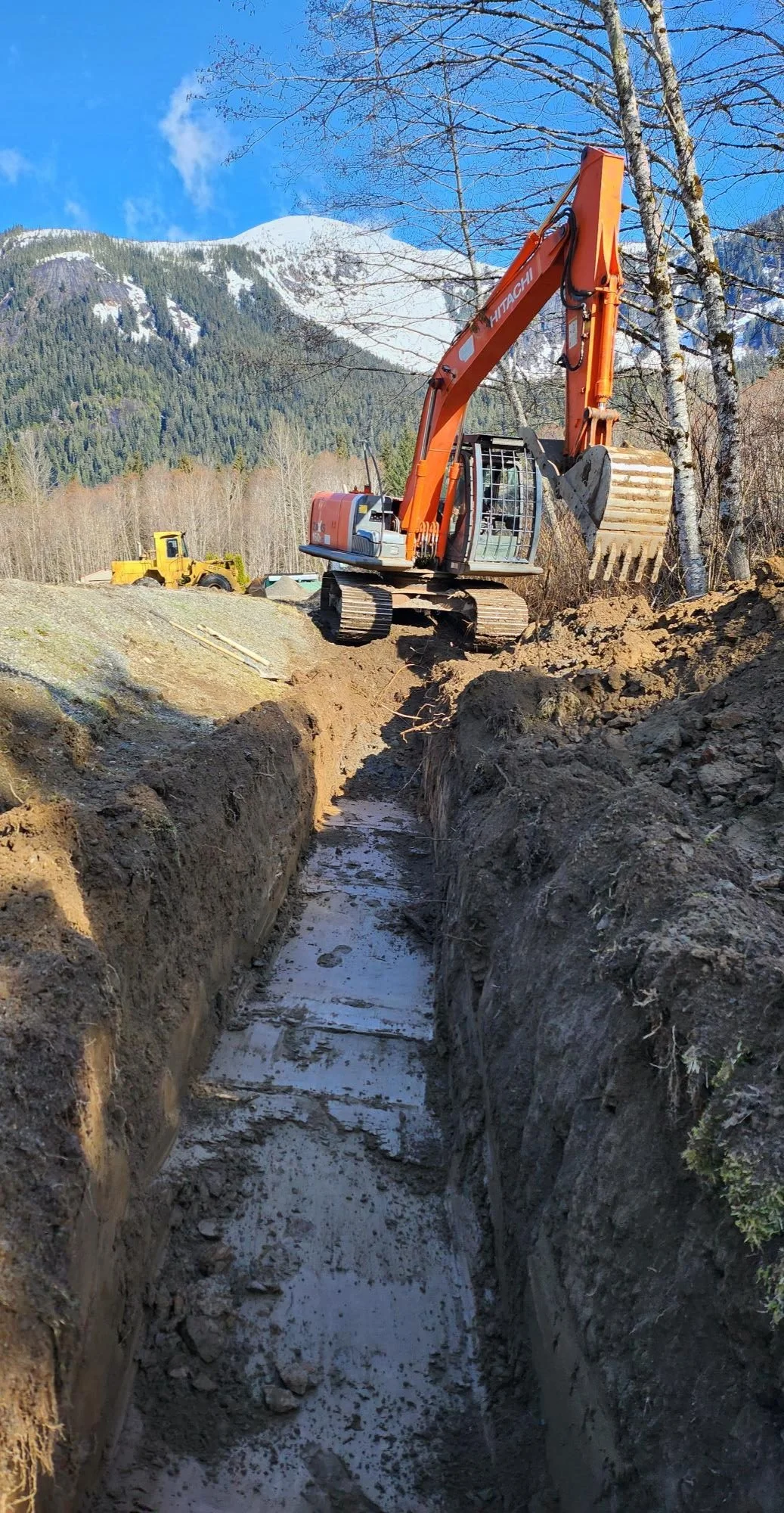 Construction site with an excavator digging a trench in a mountainous area with trees and snow in the background.