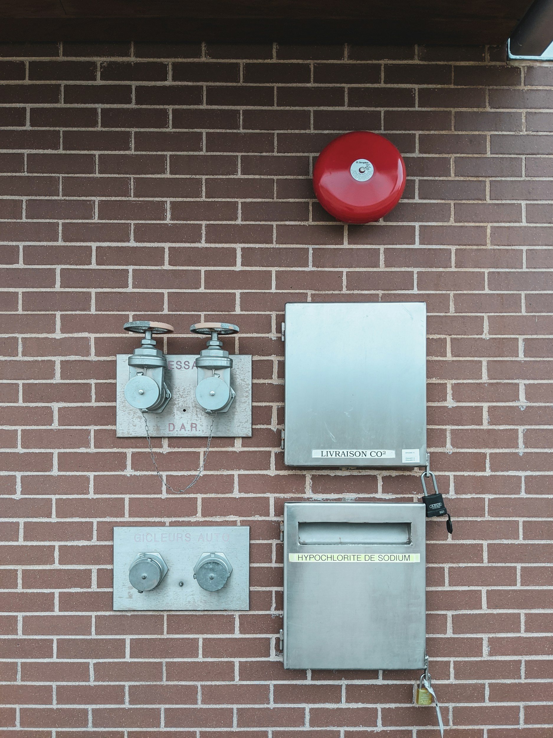 A brick wall with a red fire alarm, two grey electrical meters with chains, a grey metal box labeled 'LIVRAISON CO', and a small grey mailbox labeled 'HYPOCHLORITE DE SODIUM'.