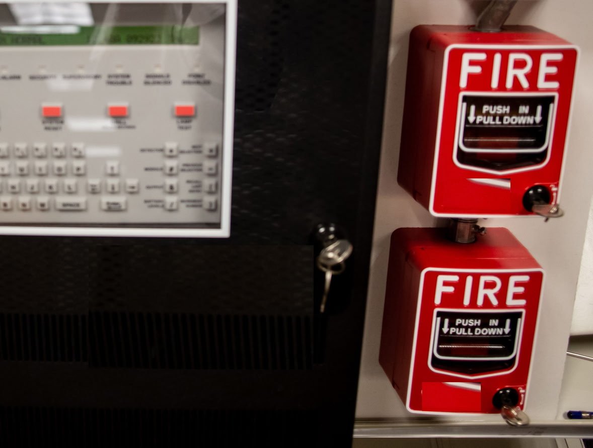 Two red fire alarm pull stations with black handles mounted on a wall next to an electronic control panel.