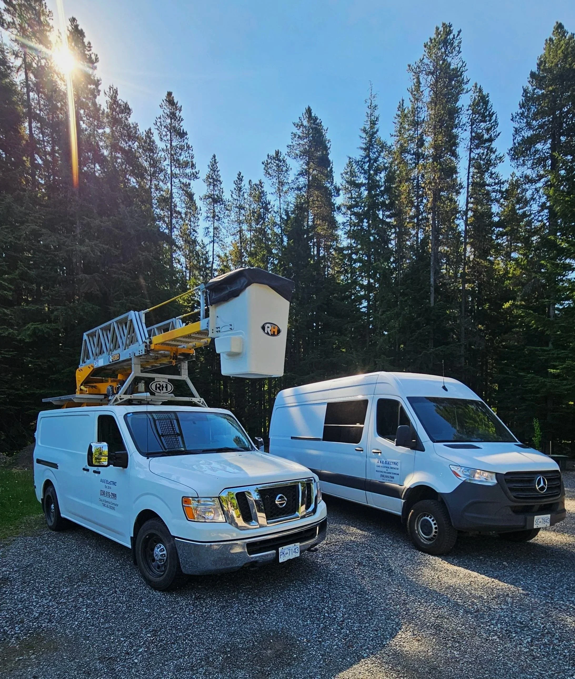 Two work trucks parked on gravel in a wooded area with tall pine trees. One truck has an aerial lift with a basket, and the other is a white cargo van, both marked with A&E Electric branding.