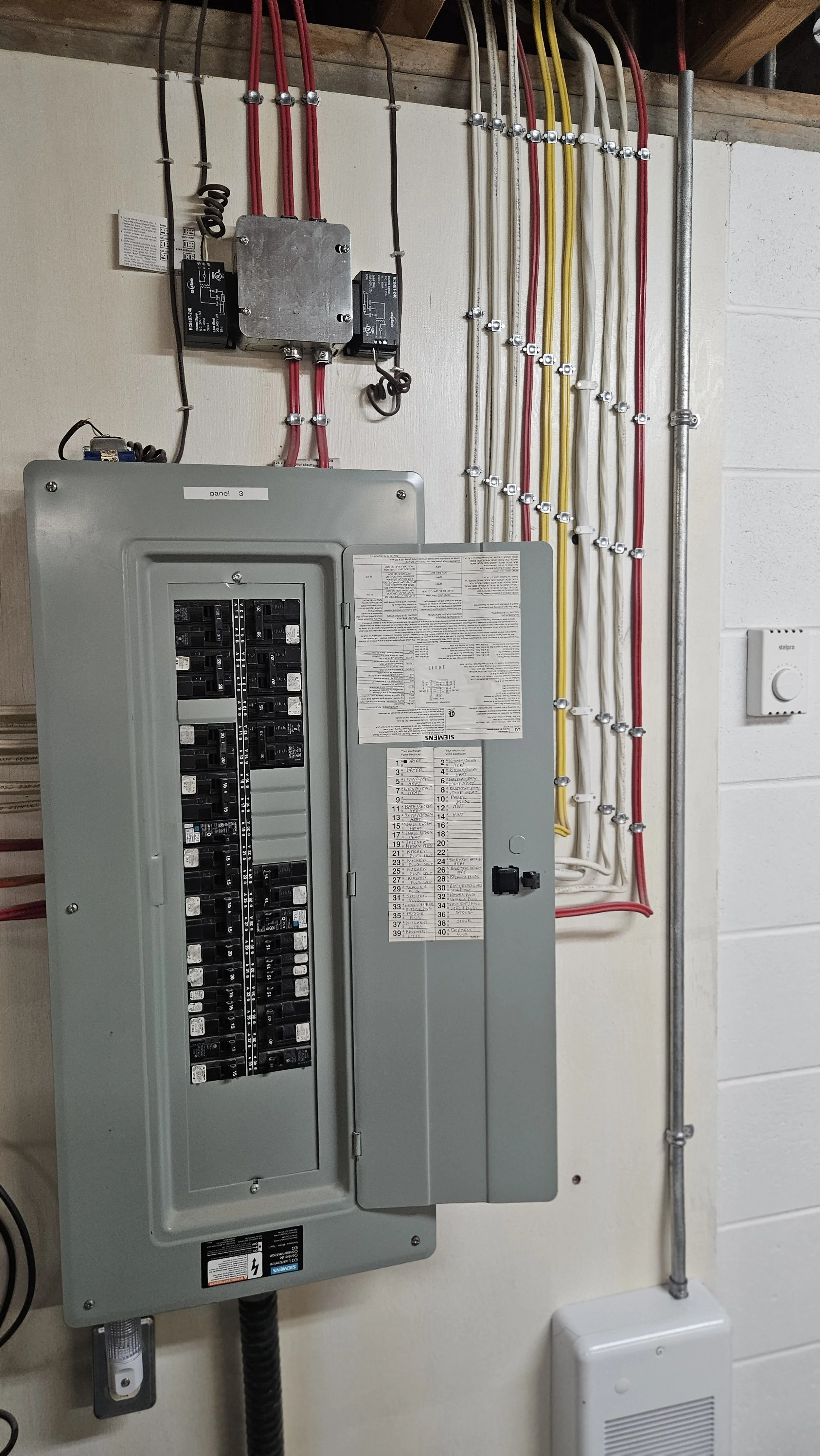 Electrical panel and circuit breakers mounted on a wall with various wires organized and secured with clips.