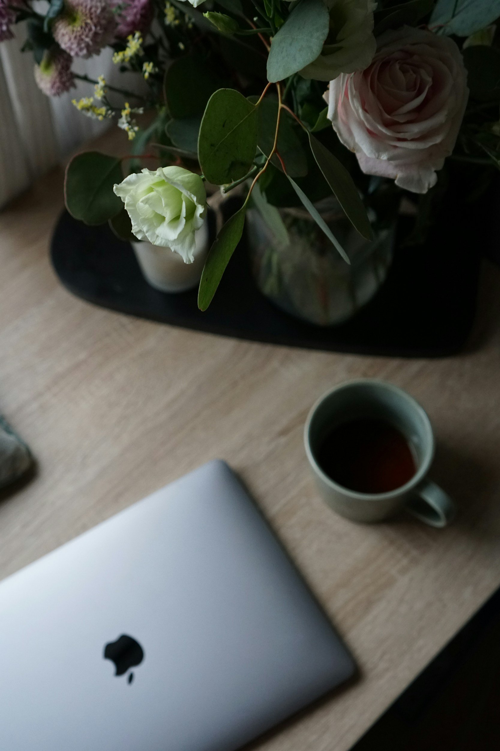 A bouquet of flowers with pink and white roses, green leaves, and small white and purple blossoms in a vase, placed on a black tray on a wooden surface. A mug with a dark liquid, a silver laptop, and a white object are also on the table.