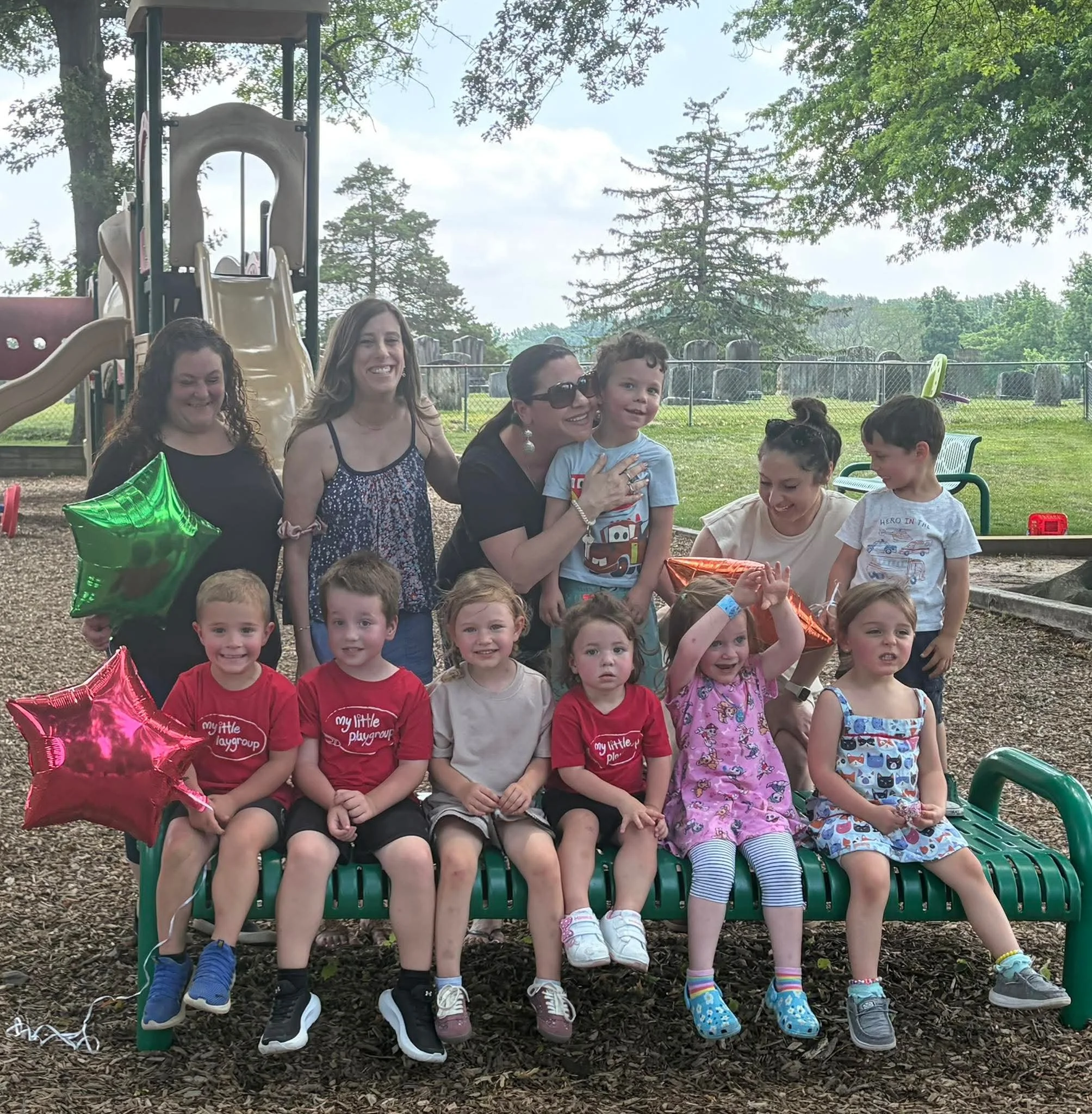 Group of children and adults sitting outdoors at a celebration with green and orange balloons, near a playground and trees.
