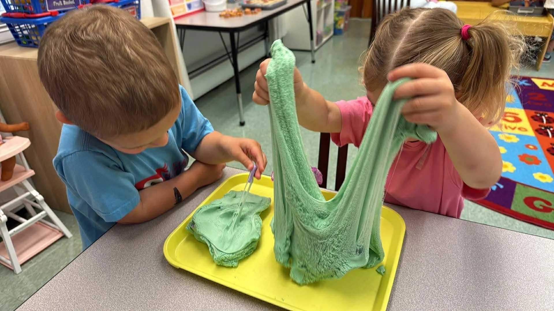 Two young children, a boy in a blue shirt and a girl in a pink shirt, are playing with green slime at a table in a classroom. The girl is stretching the slime upward, and the boy is using a small spoon to interact with the slime on a yellow tray.