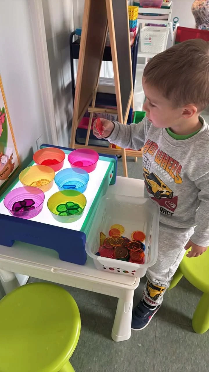 A young boy playing with translucent colored bowls and plastic buttons or tokens at a light table in a classroom or playroom.