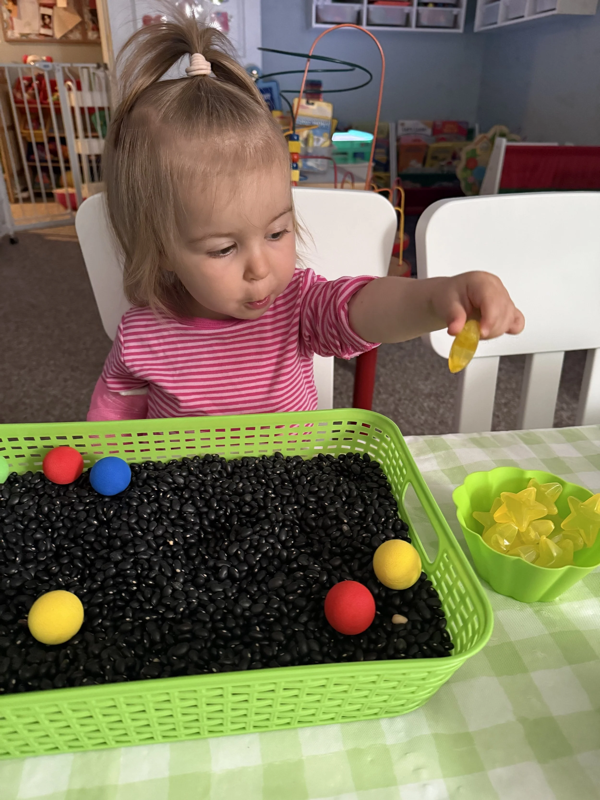 A young girl with blonde hair, wearing a pink and white striped shirt, sitting at a table in a playroom. She is holding a yellow toy in her right hand and looking at a green basket filled with black beans, with red, yellow, and blue foam balls scatte