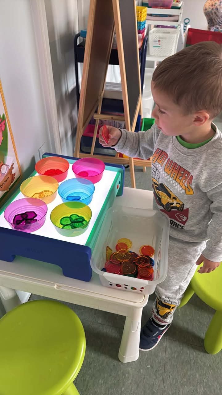 A young boy playing with colorful translucent cookies and a stacking toy on a light table in a classroom or playroom.
