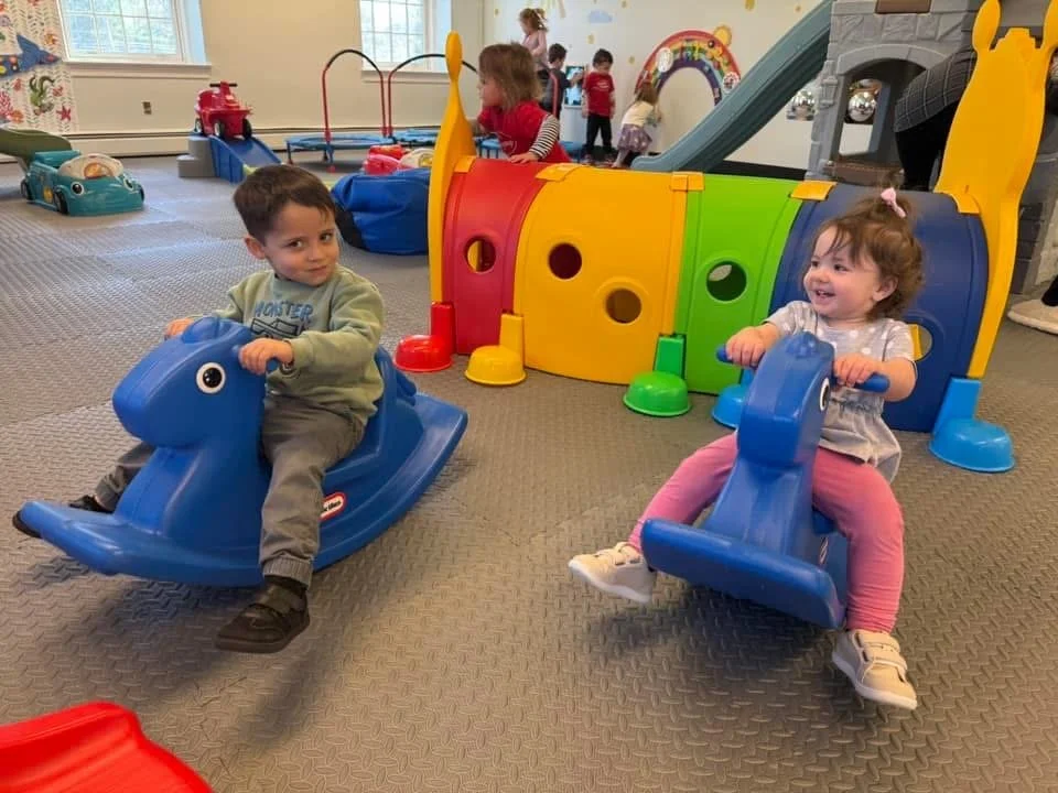 Two young children playing on blue animal-shaped rocking toys in an indoor play area, with other kids and colorful play structures in the background.
