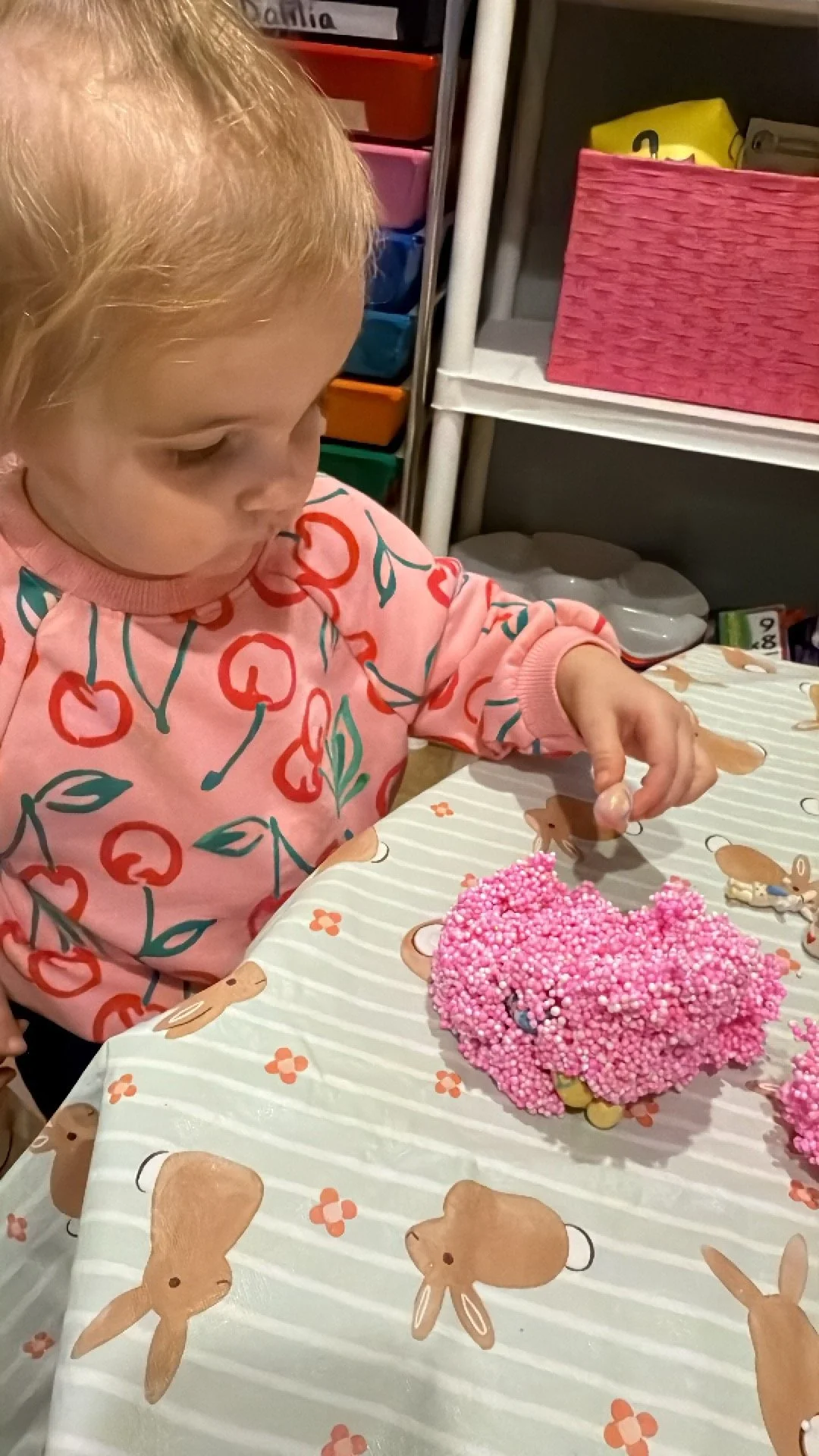 A young girl with blonde hair wearing a pink shirt with cherries, sitting at a table decorated with bunny and flower patterns, looking at a pink beaded cake.