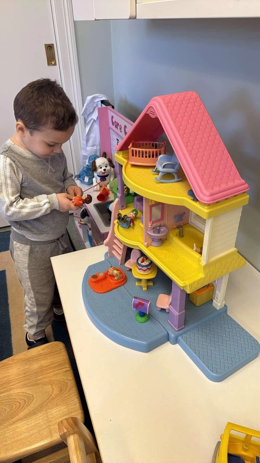 A young boy playing with a large plastic dollhouse on a table surrounded by chairs, with various toy accessories and figures.