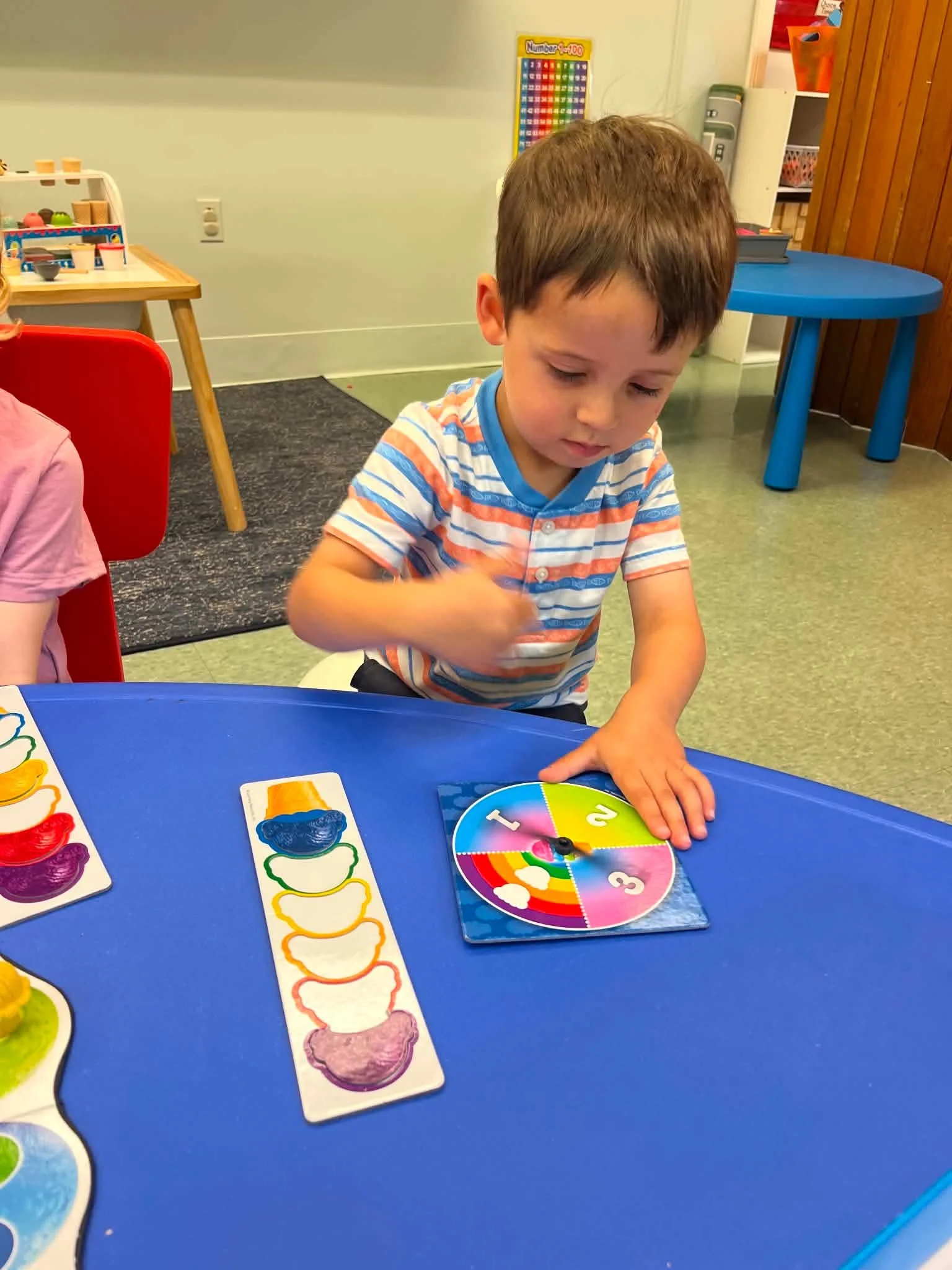 A young boy with brown hair wearing a striped shirt playing a colorful game on a blue table in a classroom setting, with educational posters and toys in the background.
