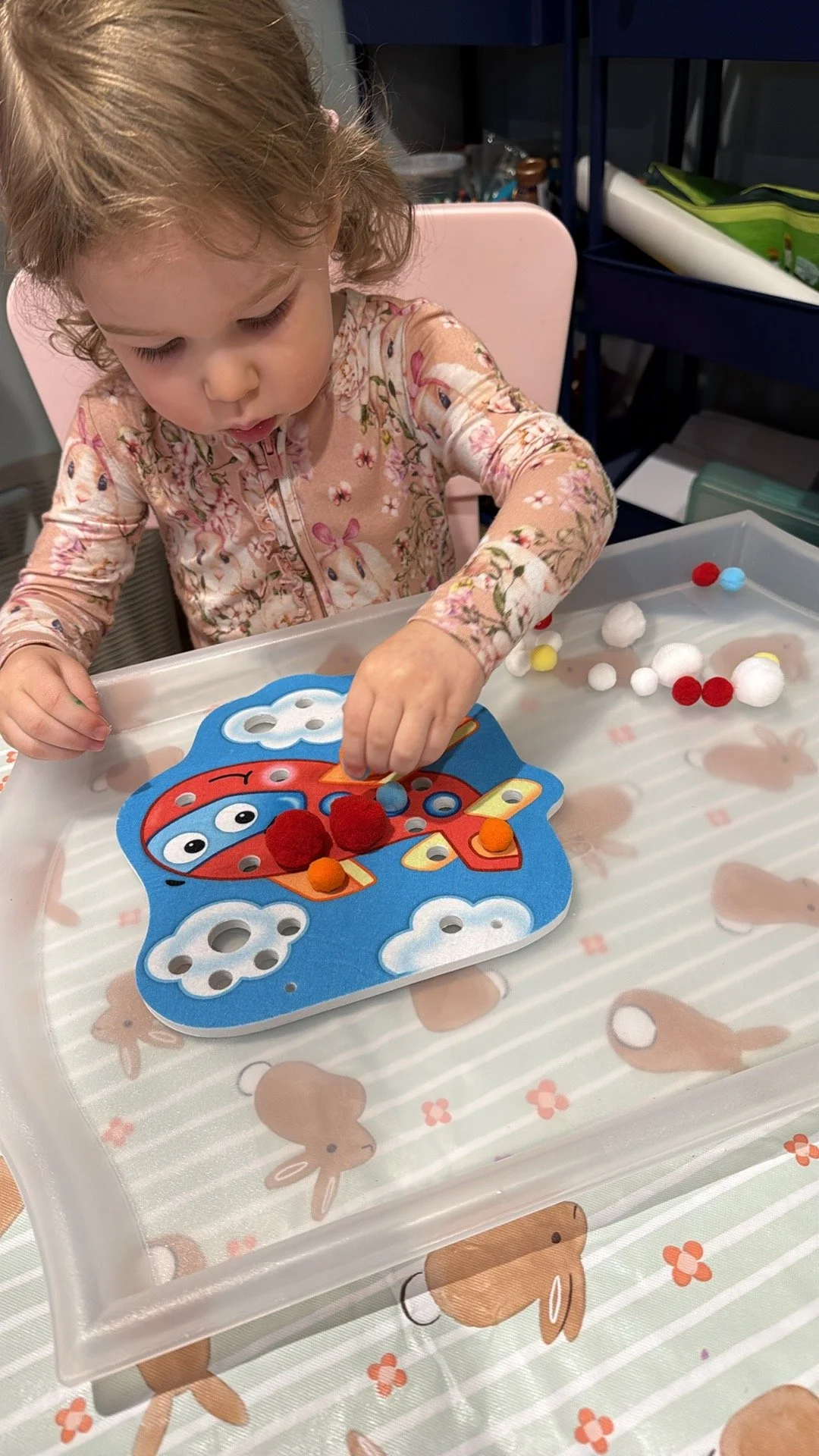 A young girl in bunny pajamas plays a game with a colorful ladybug-shaped board on a table with bunny designs, using small fuzzy and spherical game pieces.