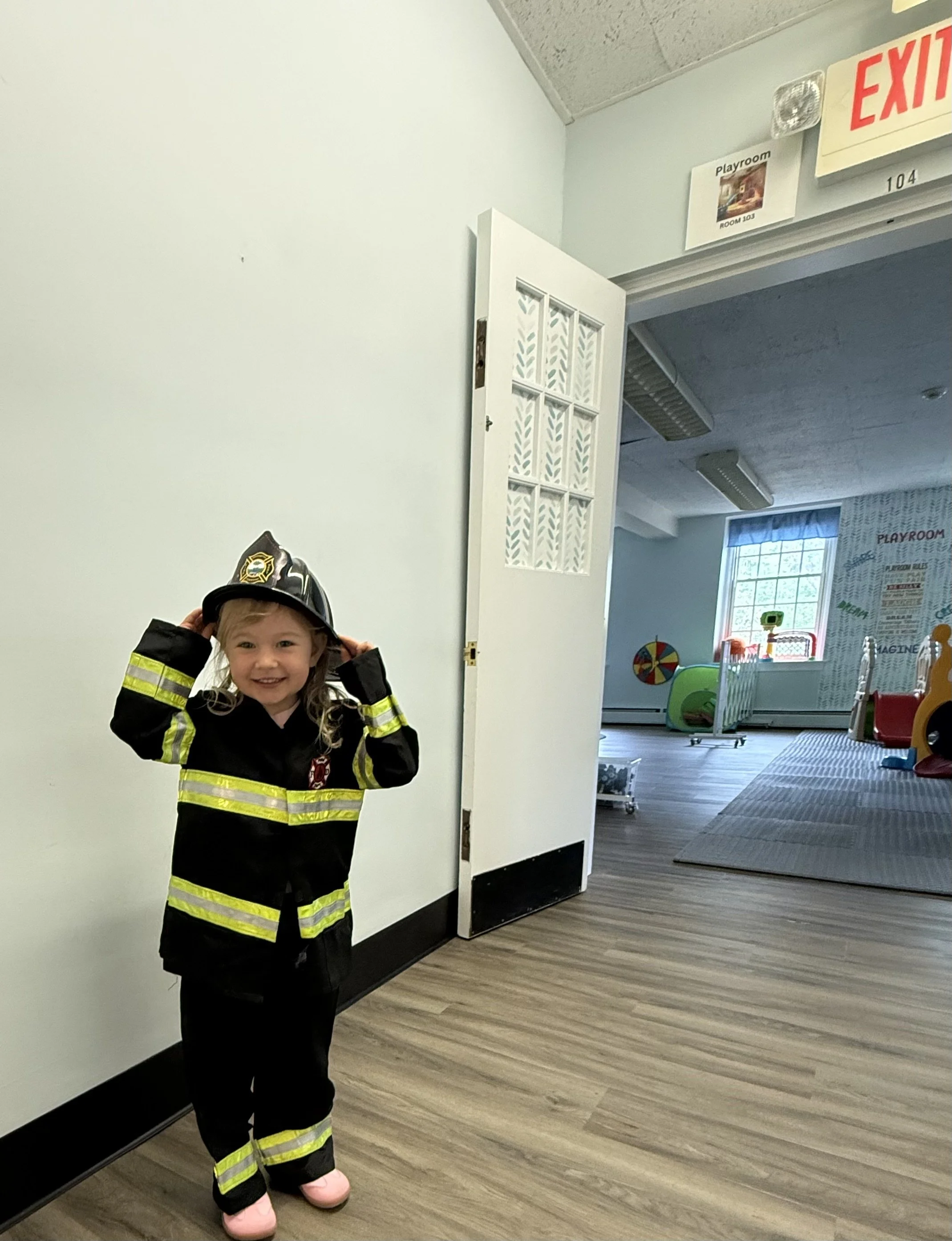 Young girl dressed as a firefighter in a toy playroom, smiling and holding her firefighter hat, with colorful toys and play area visible in the background.