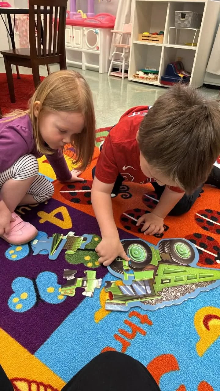 Two children, a girl with blonde hair and a boy with brown hair, are putting together a puzzle on a colorful rug in a playroom. The puzzle depicts a green and black robot or vehicle. The background shows furniture, including a dollhouse, a white book