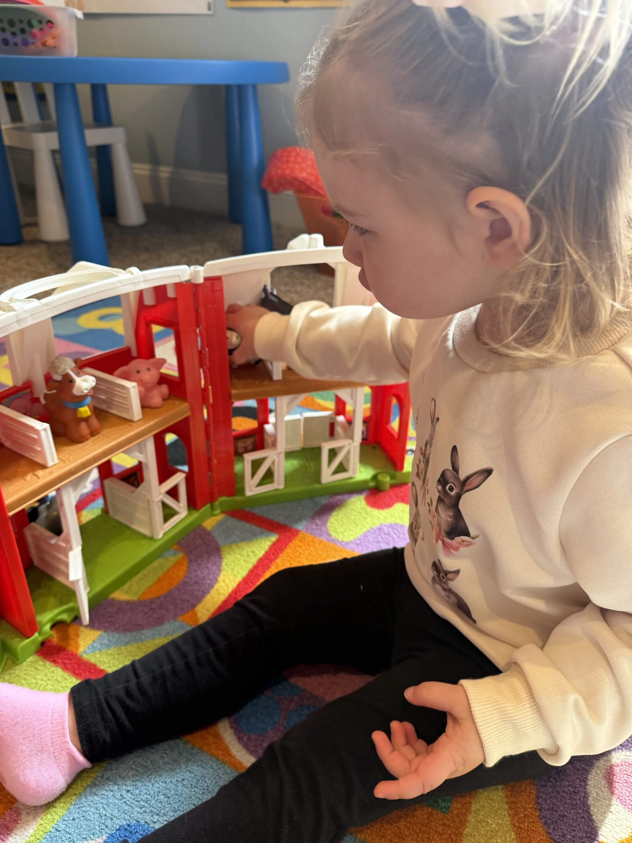 A young girl is playing with a toy barn and animals on a colorful rug in a playroom.