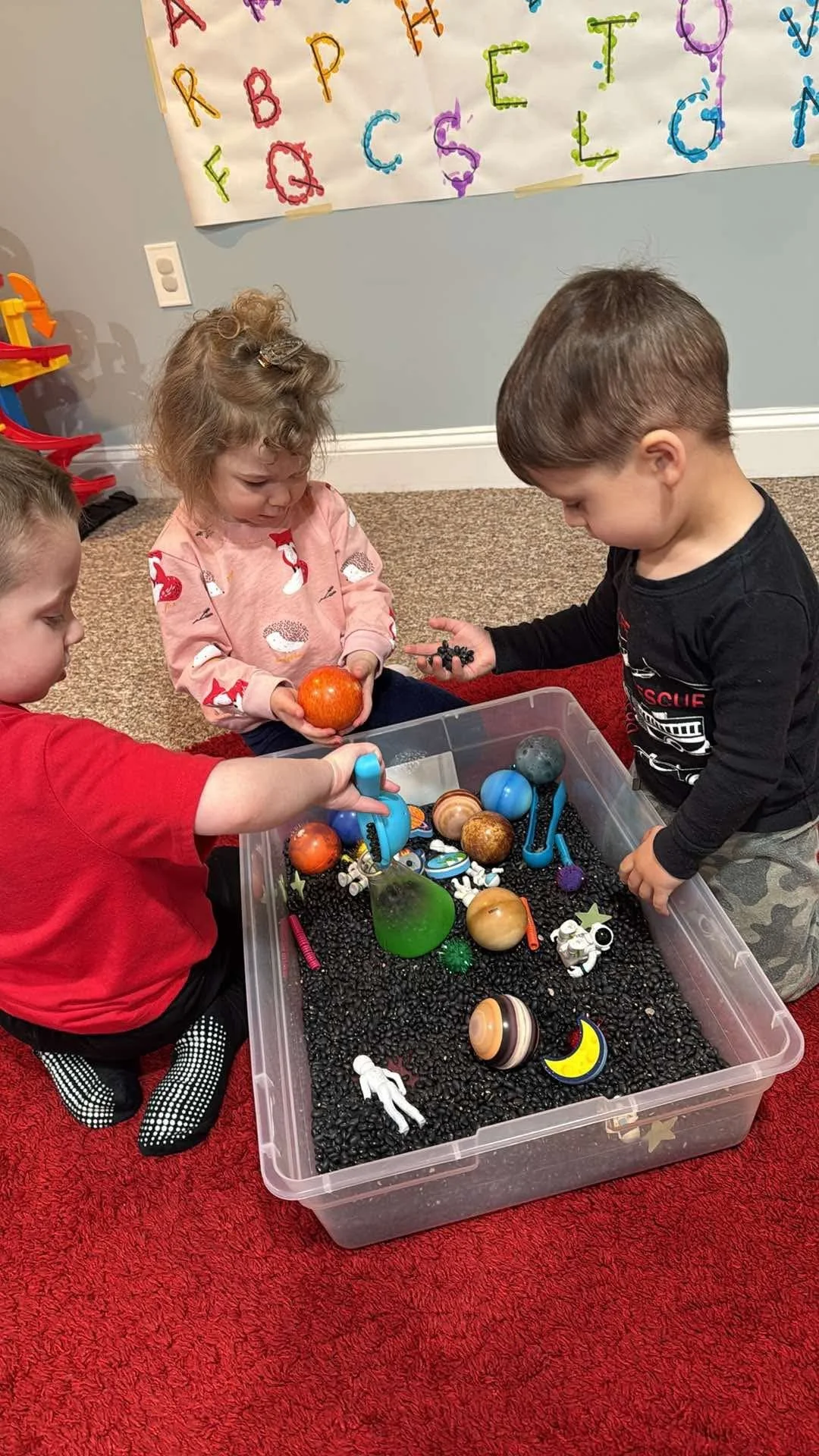 Three children with light skin playing with space-themed toys in a plastic bin filled with small black beads, set on a red carpet, with a poster of the alphabet on the wall behind them.