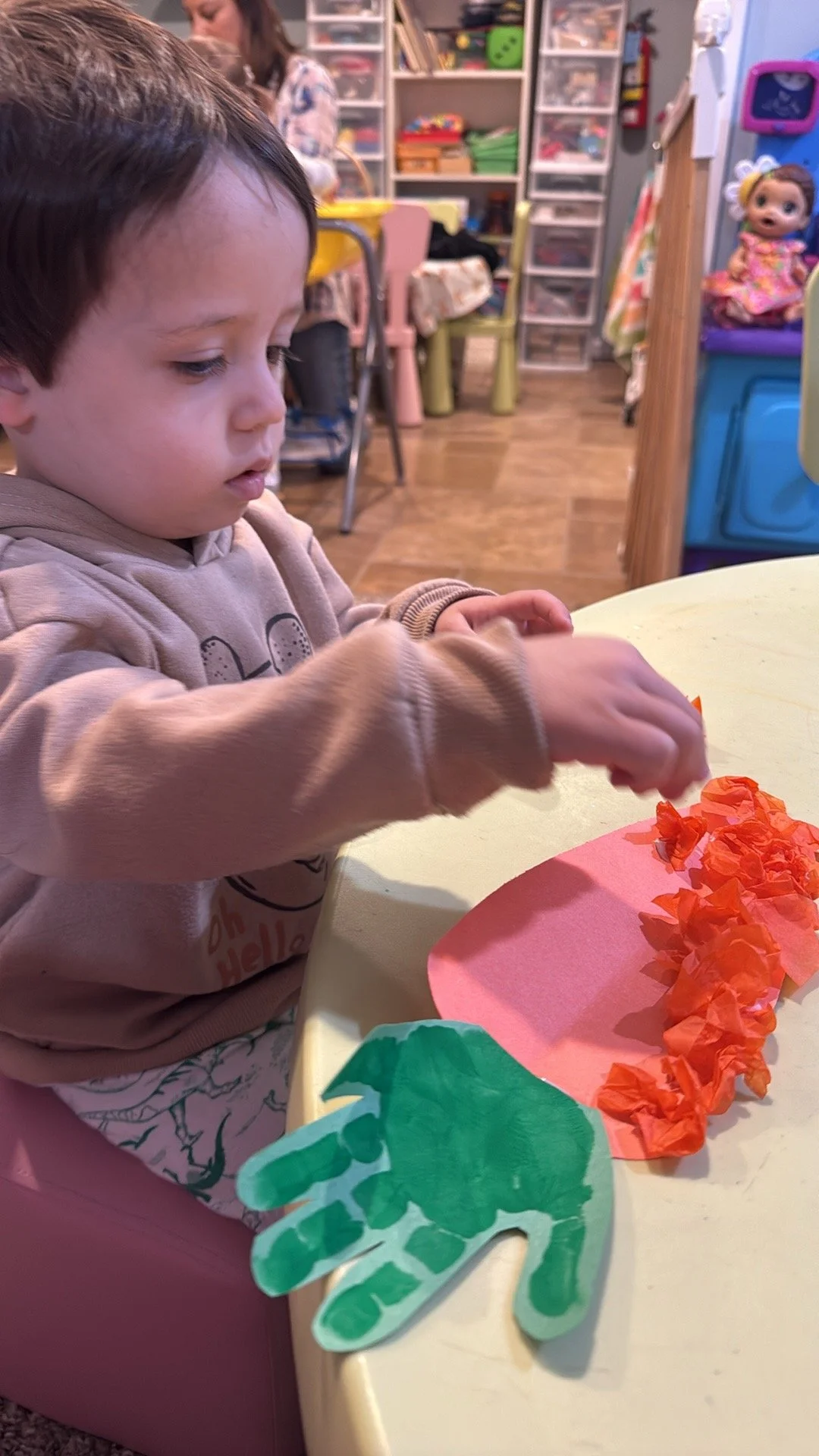 A young child sitting at a yellow table, decorating a paper craft with orange tissue paper, in a colorful and cluttered craft room.