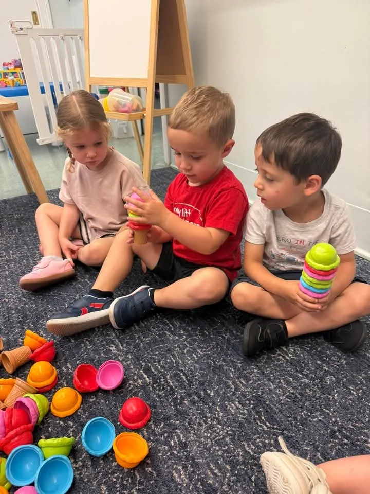 Three children sitting on the carpeted floor with colorful stacking cups and toy fruits nearby, in a playroom or classroom setting.