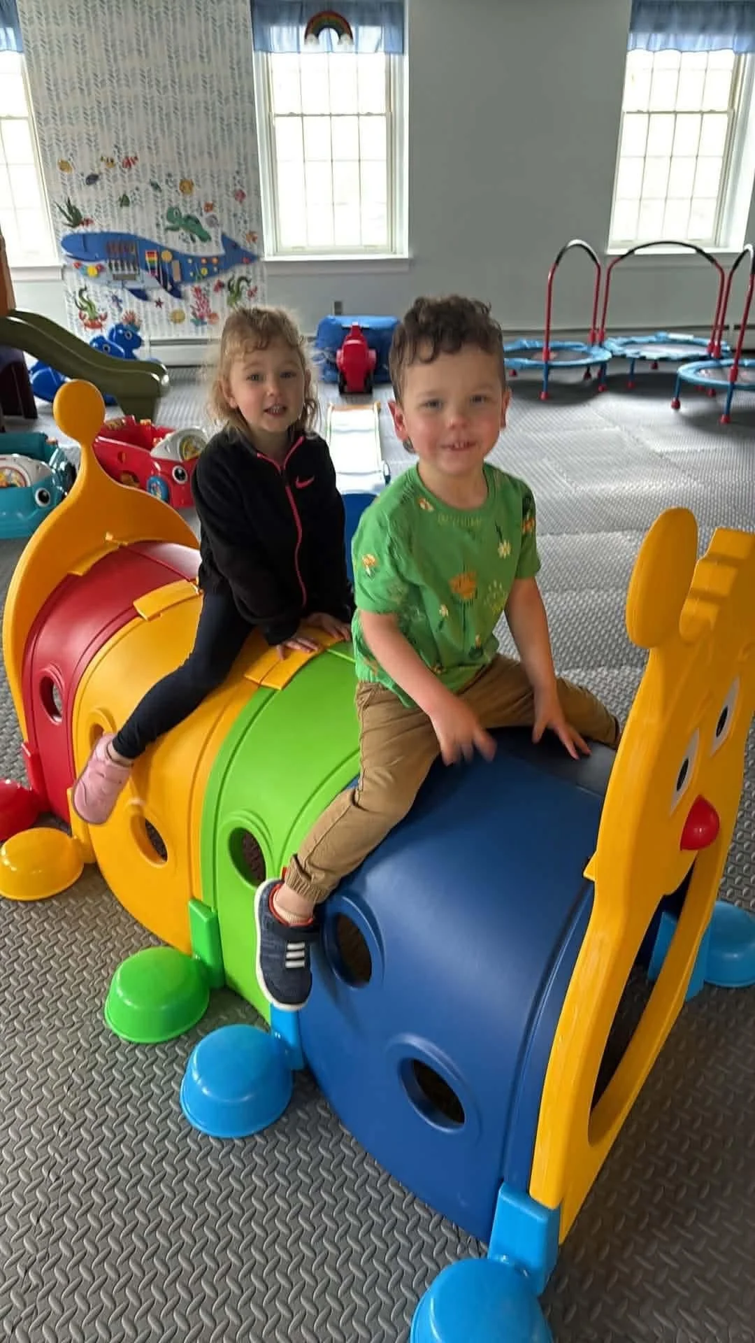 Two children, a girl and a boy, playing on a colorful plastic toy in an indoor play area with windows in the background.