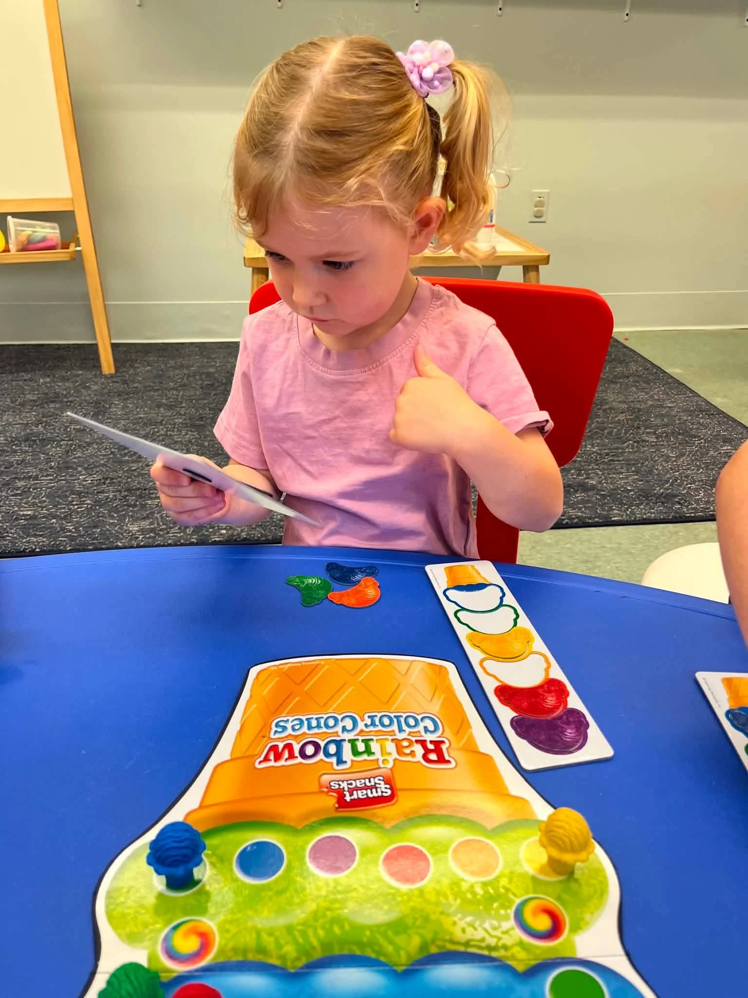 A young girl with blond hair tied with a purple scrunchie, sitting at a blue table, looking at a tablet or phone in her hand, with a colorful coloring game and a rainbow colors paint set on the table in front of her.