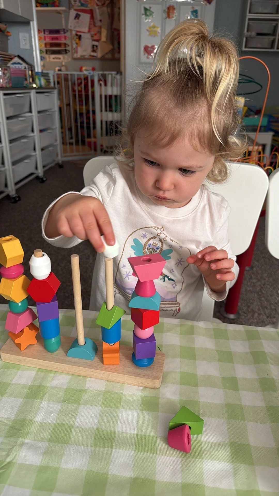 A young girl with blonde hair styled in a high ponytail is sitting at a table, stacking colorful wooden blocks on wooden sticks as part of a toy activity. The background shows a playroom with toys, shelves, and a playpen.