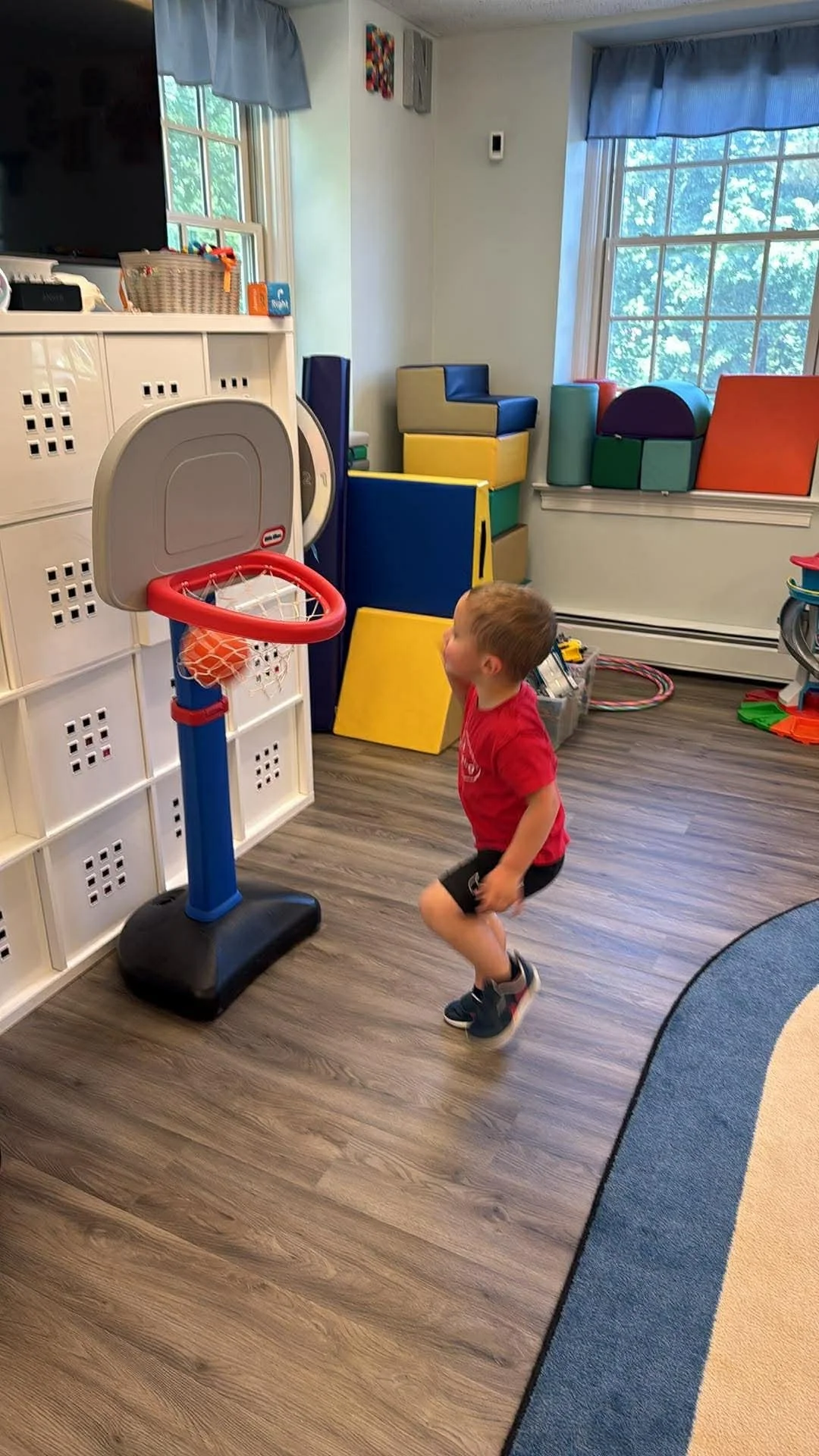 A young boy in a red shirt and shorts playing basketball indoors with a small basketball hoop.