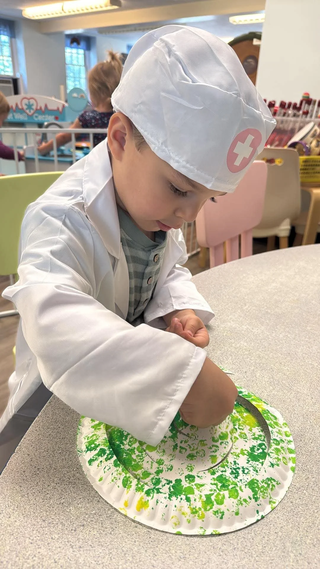 A young boy dressed as a doctor, wearing a white coat and a white hat with a red cross, is engaged in a craft activity on a table, using green and yellow paint on a paper plate with a green spiral design. The background shows a classroom setting with