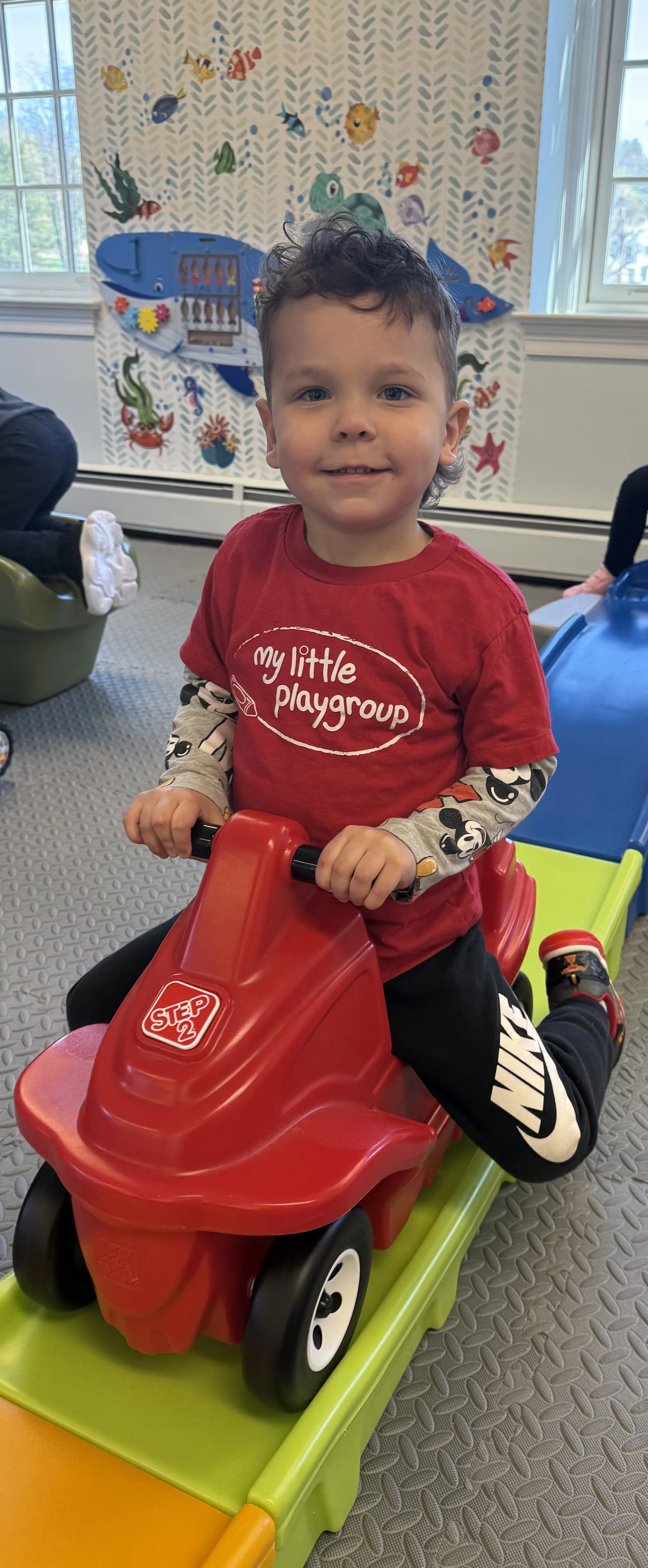 A young boy with curly hair riding a red and green plastic toy car in an indoor play area, wearing a red shirt that says 'my little playgroup' and black pants.